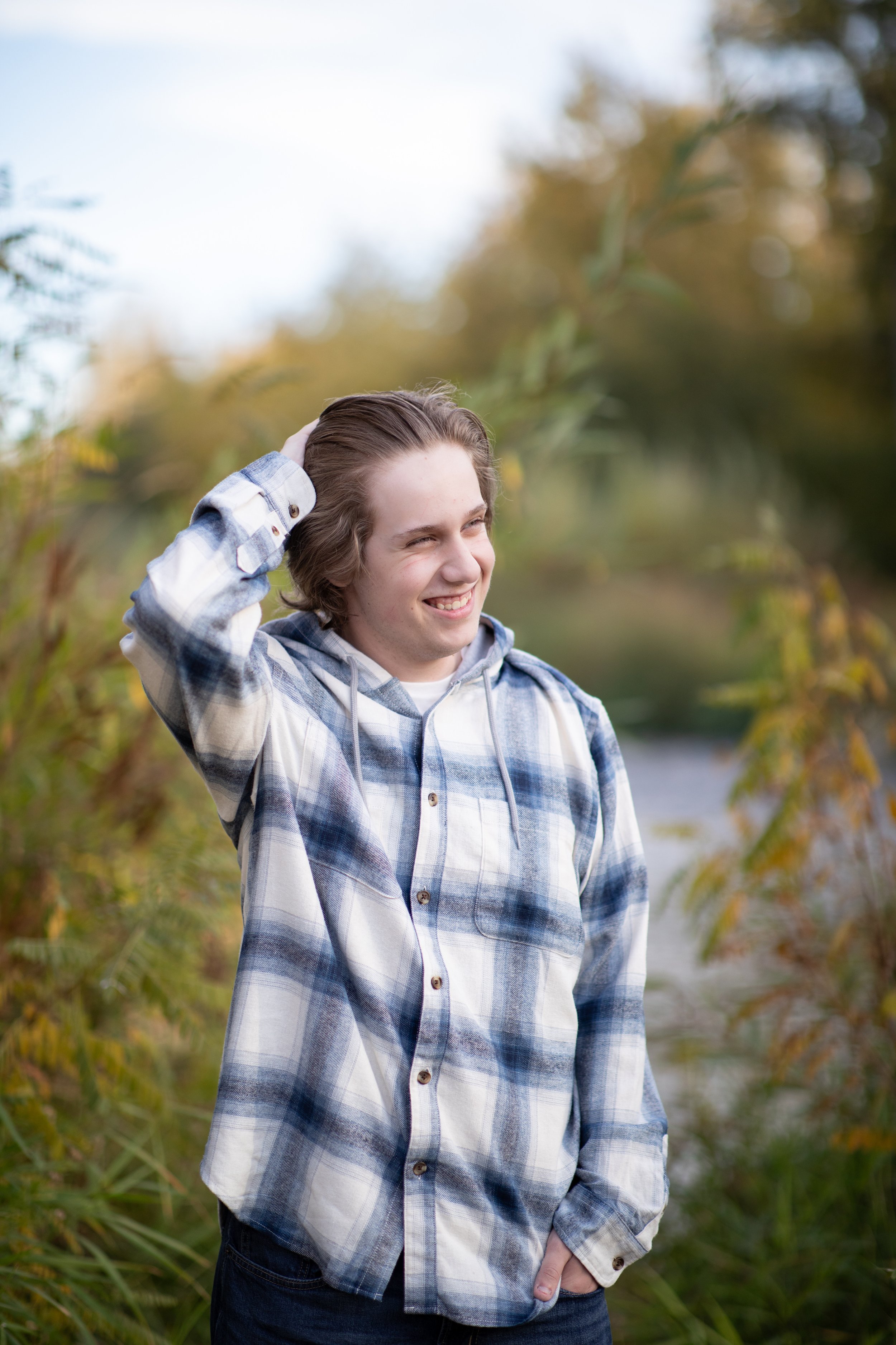 A young man outdoors by a river, smiling and touching his hair, wearing a plaid shirt with fall foliage in the background.