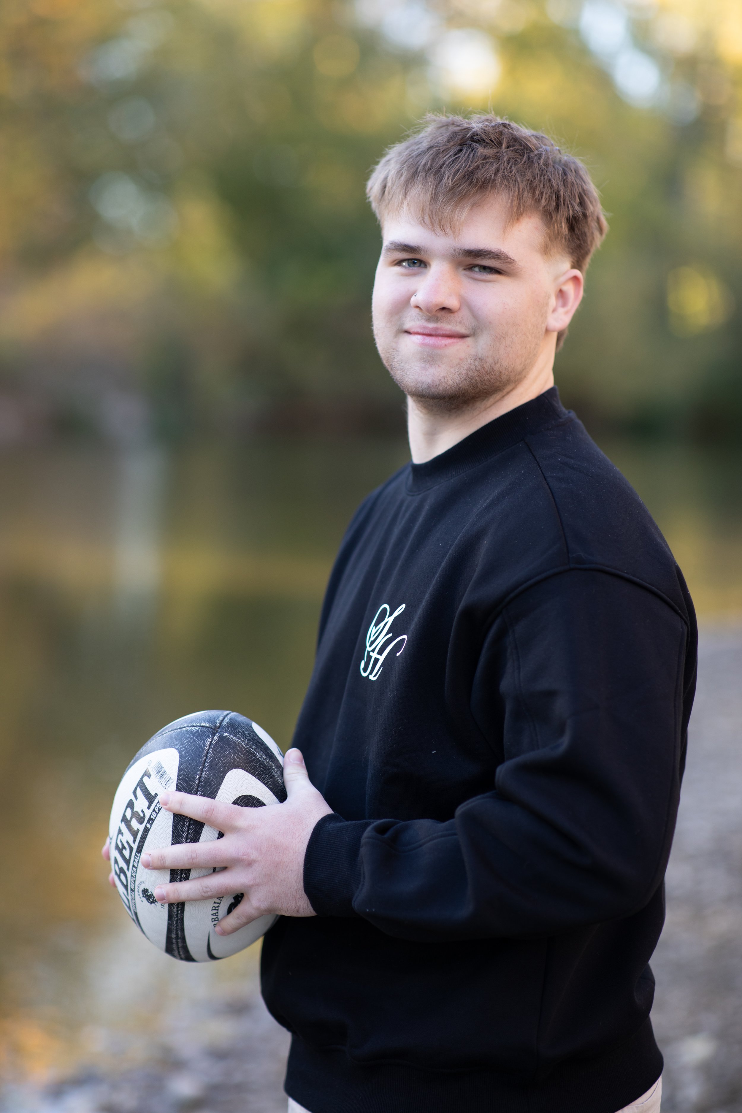 Young man with light brown hair, wearing a black sweatshirt with a monogram logo, holding a rugby ball, outdoors near water with blurred trees in the background.
