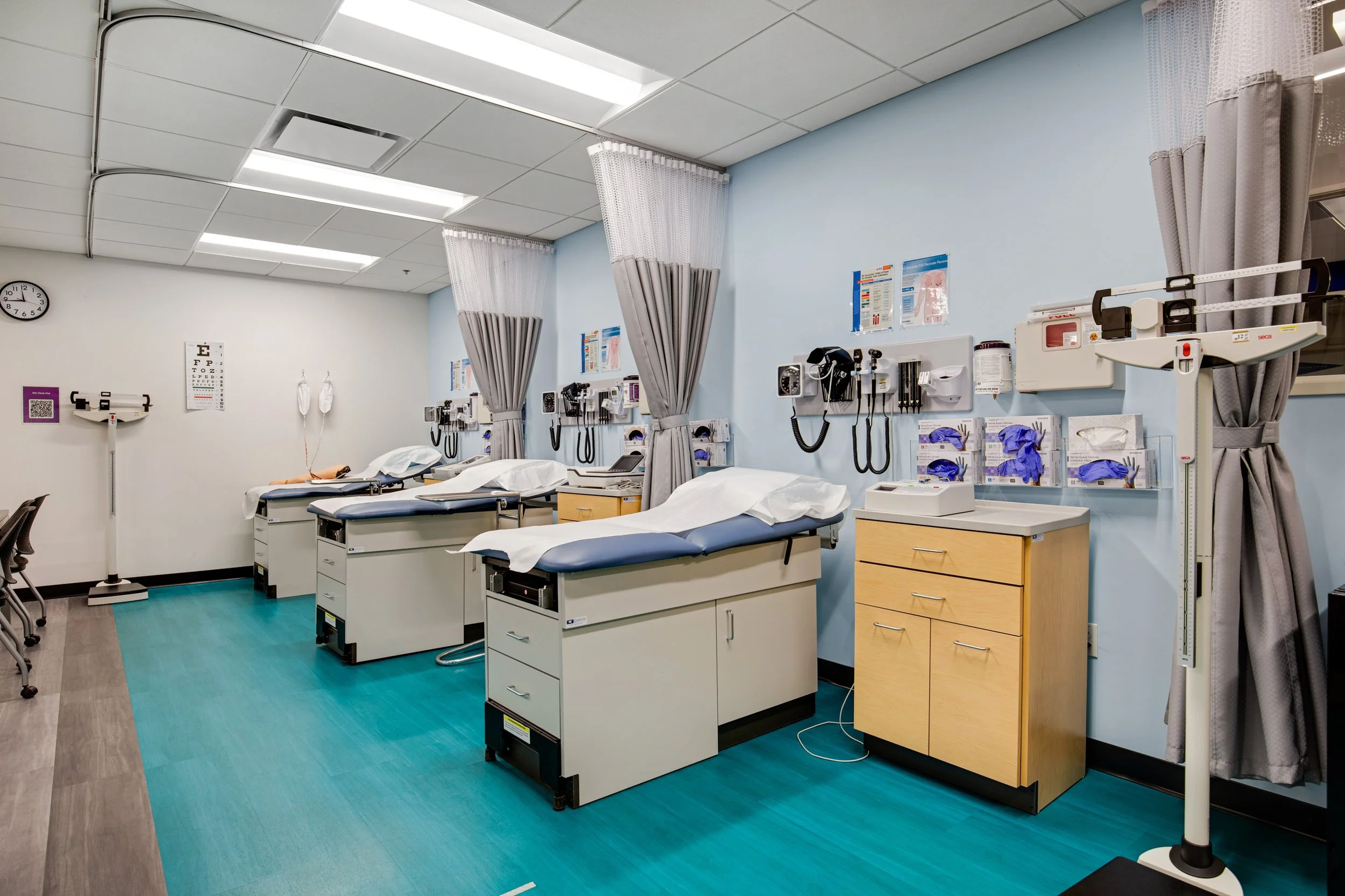 Empty hospital or clinic examination room with three patient beds, each with medical equipment, curtains separating the beds, and medical charts and supplies on the walls.