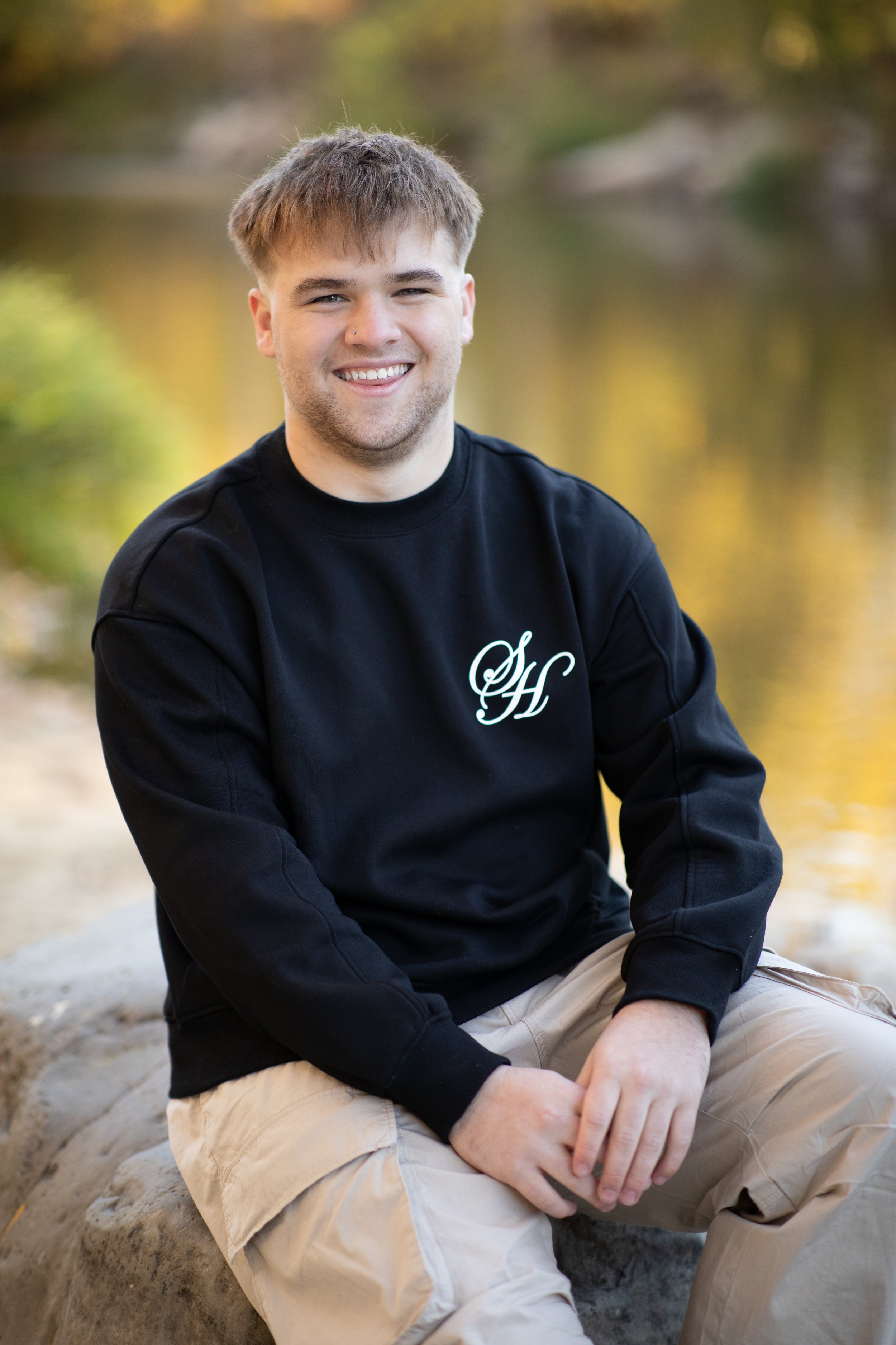 Young man sitting outdoors on a rock, smiling, with a blurred natural background including a body of water and trees.