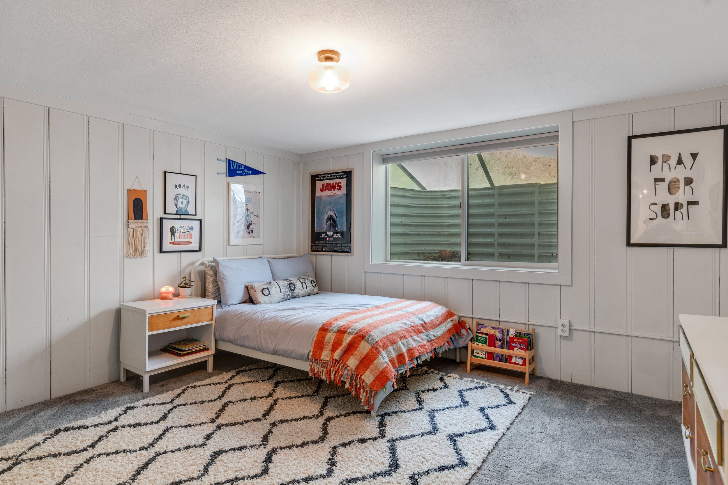 A bedroom with white wooden paneled walls, a bed with gray bedding and a striped orange and white blanket, a side table with a small lamp, and several posters and framed artworks on the wall, including a Jaws poster and a "Pray for Surf" print. There
