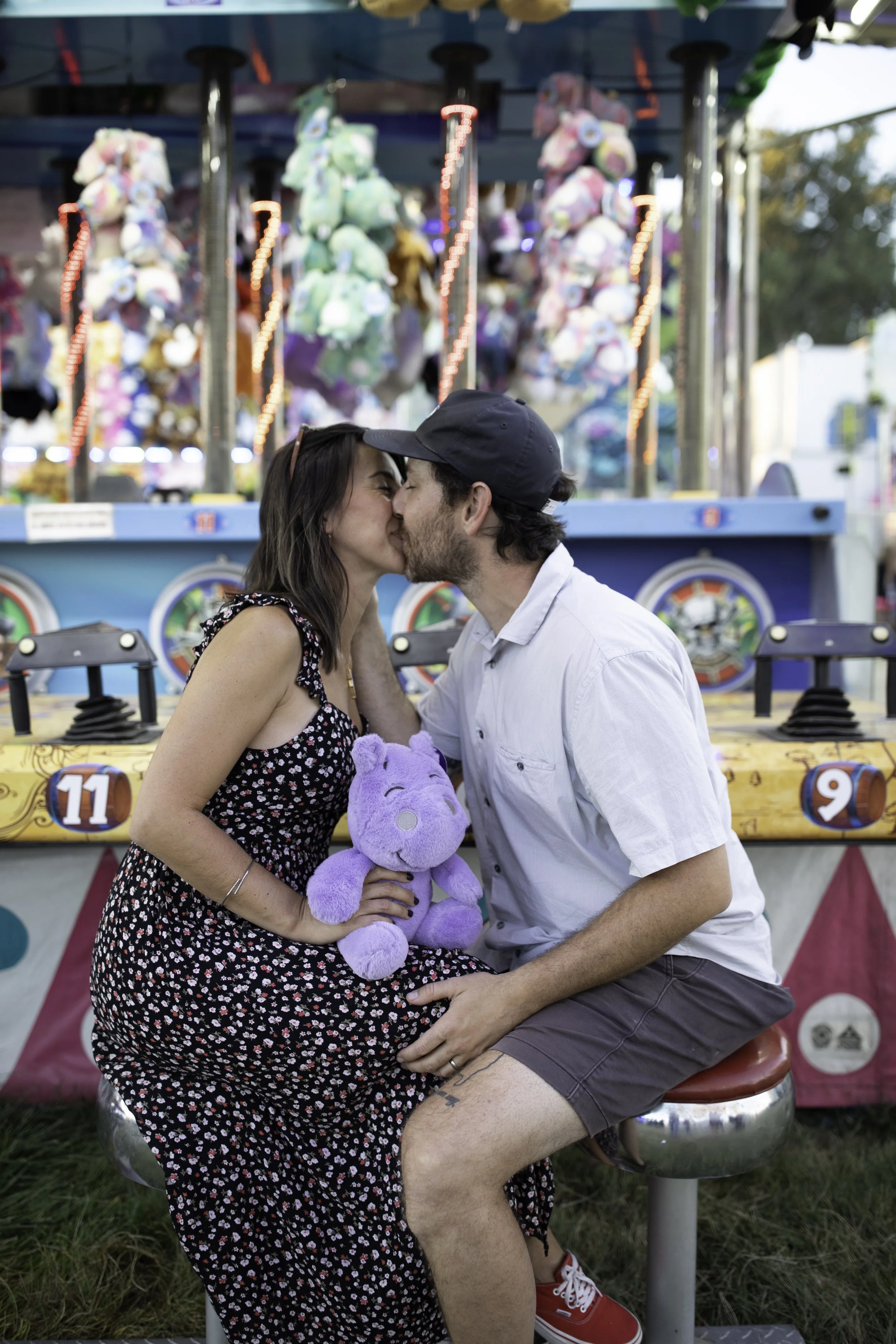 A couple kissing at a carnival, with a plush purple hippo toy, in front of game booths with plush toys and bright lights.