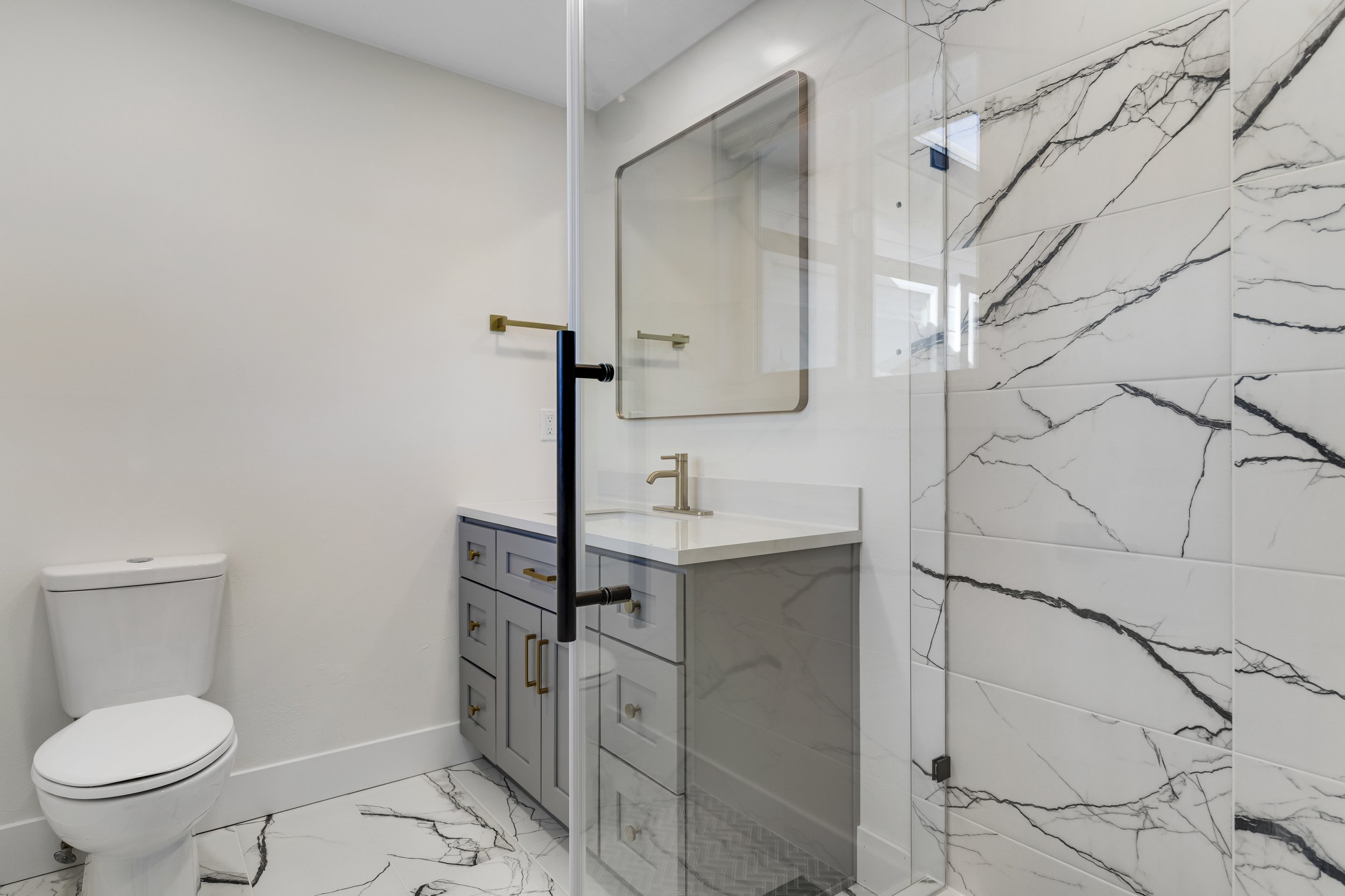 Modern bathroom with white walls, marble tiles, a toilet, a gray vanity with gold hardware, a rectangular mirror, and a glass shower enclosure with black accents.