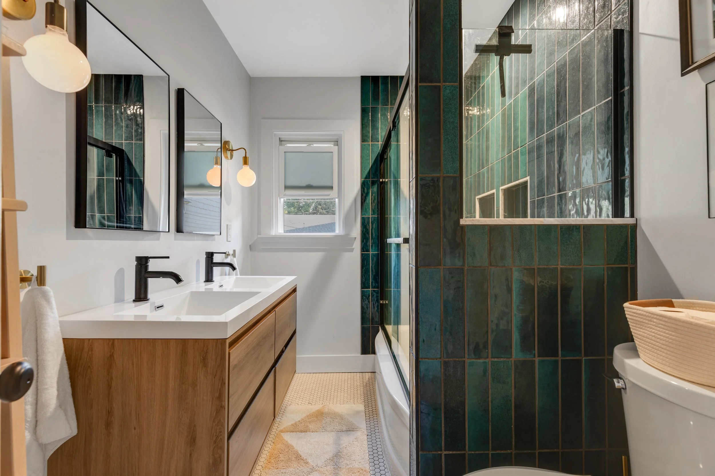 Modern bathroom with dual sink vanity, black faucets, large mirrors, wall-mounted lights, green tiled walk-in shower, and white toilet with wicker basket. Small window with shades.