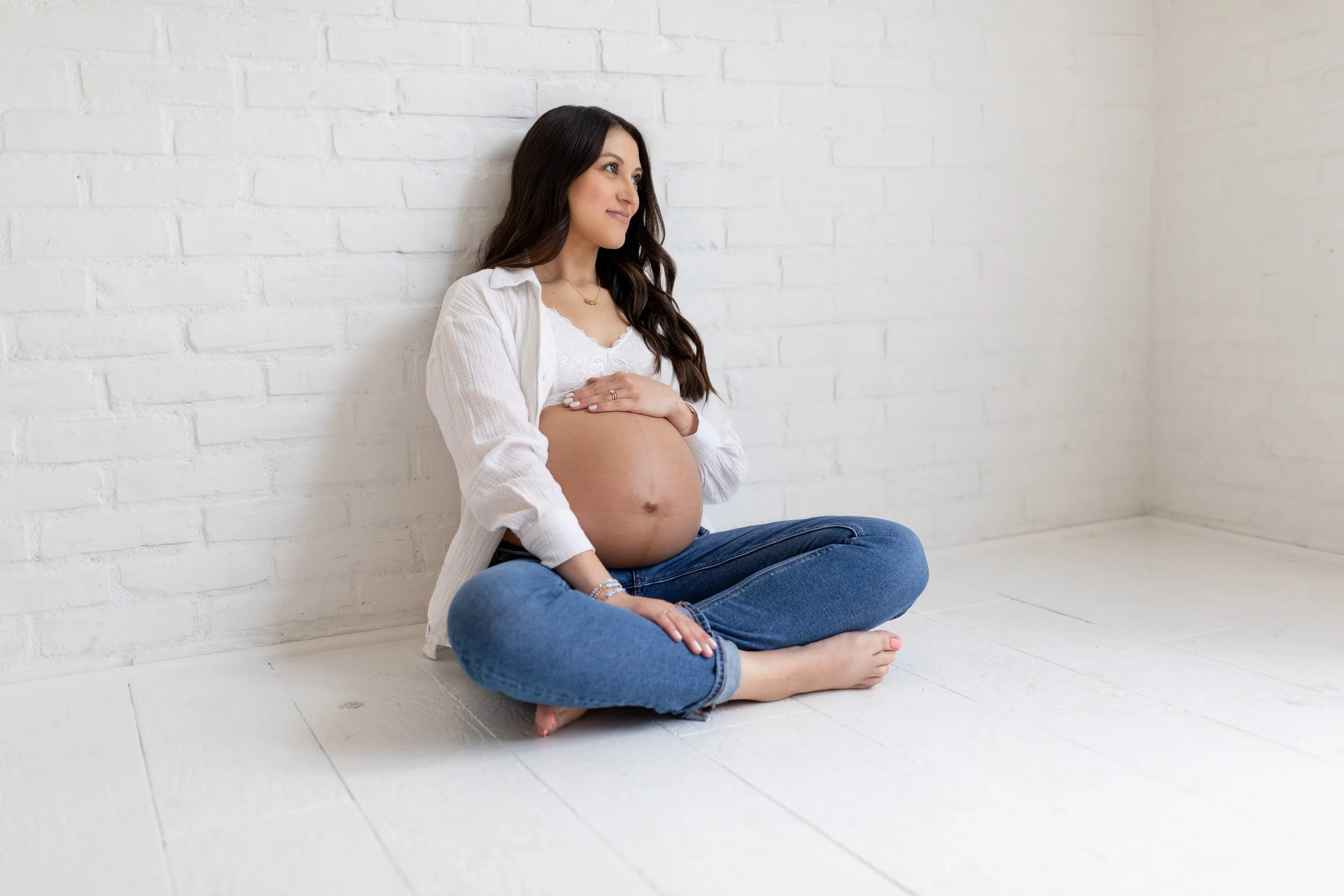 Pregnant woman sitting cross-legged on white floor, leaning against white brick wall, wearing jeans and a white top, with her hands resting on her belly.