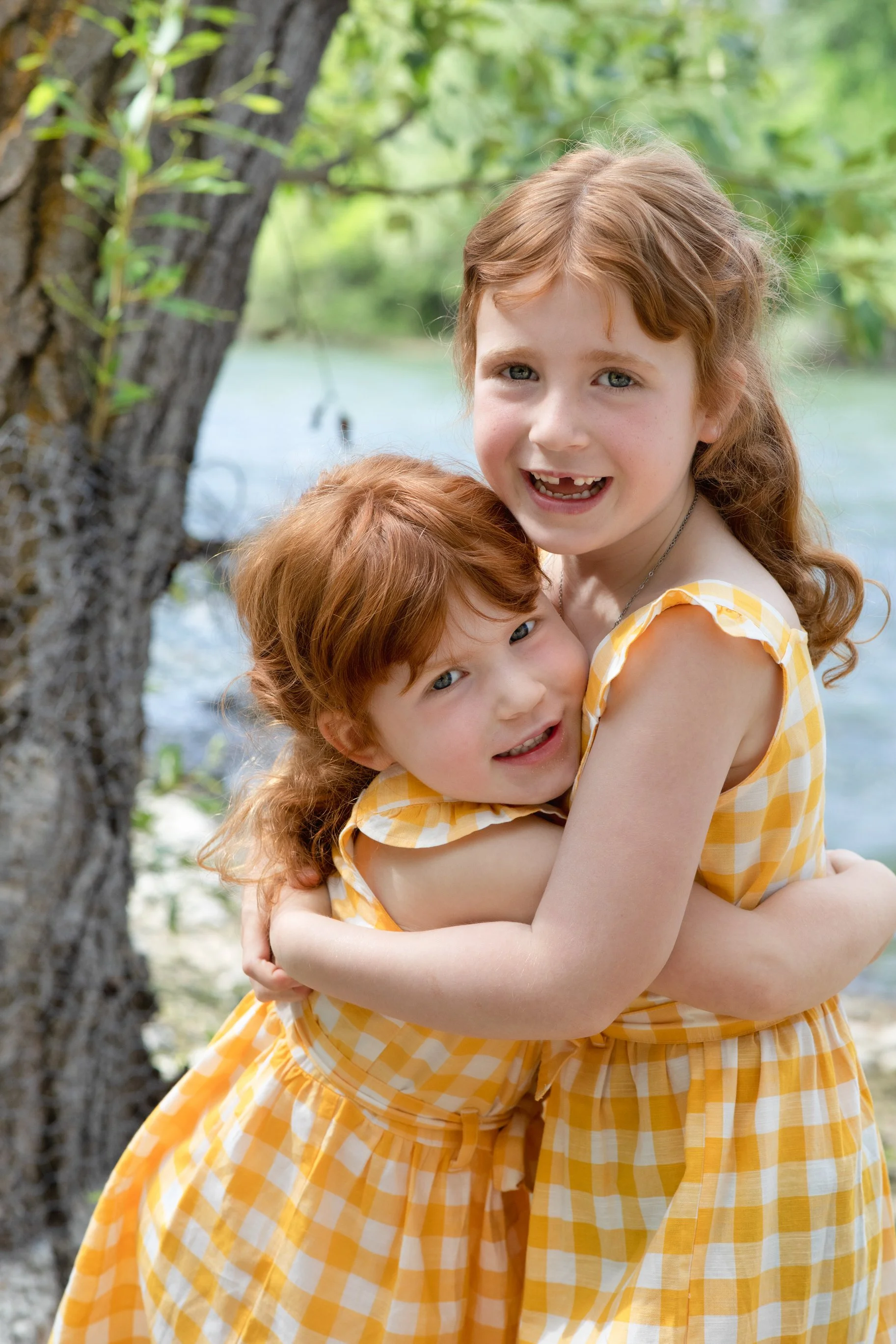 Two girls with red hair, wearing yellow gingham dresses, hugging each other outdoors near a tree and water.