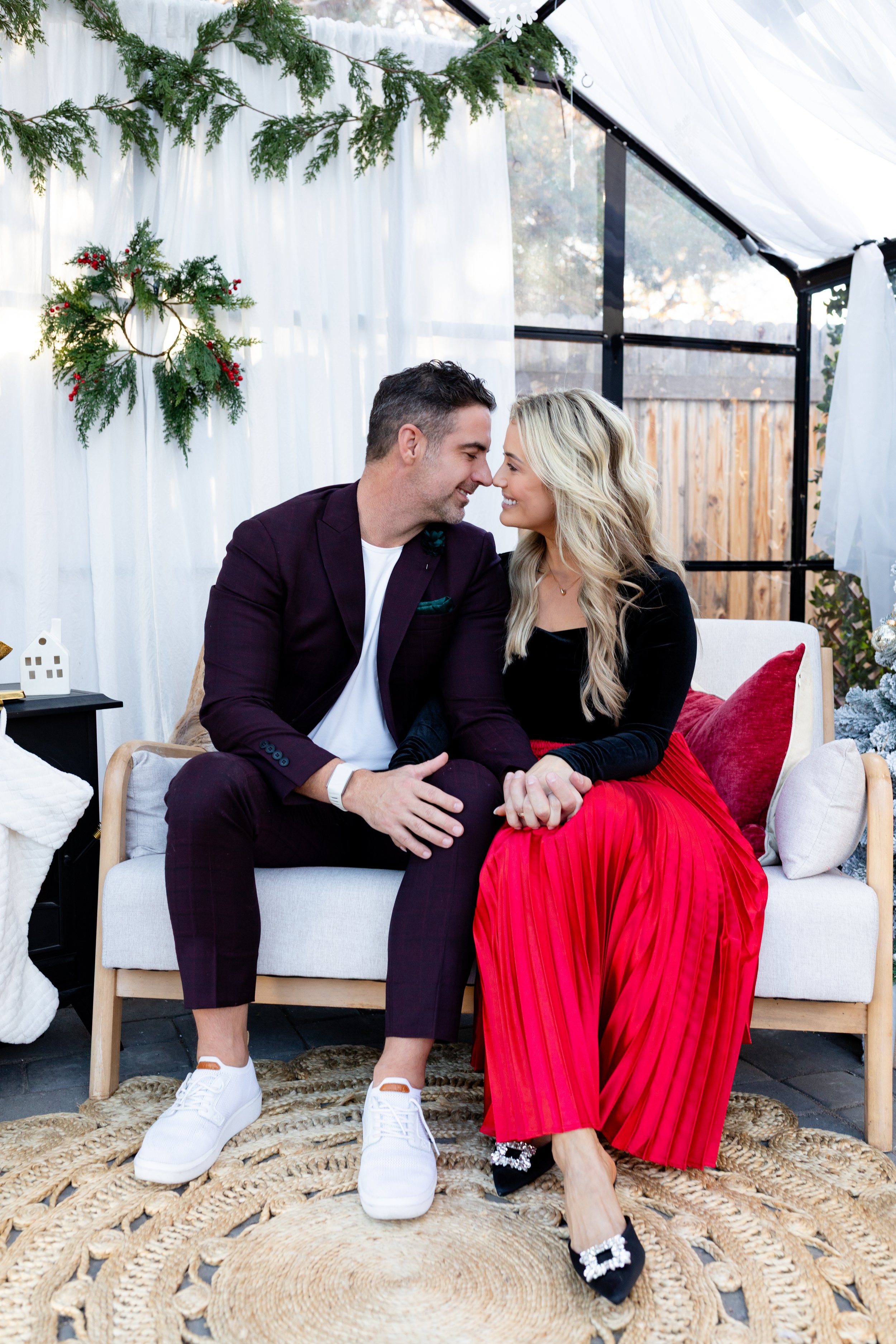 A man and woman sitting close together on a white sofa, holding hands and smiling affectionately at each other, in a decorated indoor space with holiday decorations like a small Christmas tree, wreath, and white curtains.