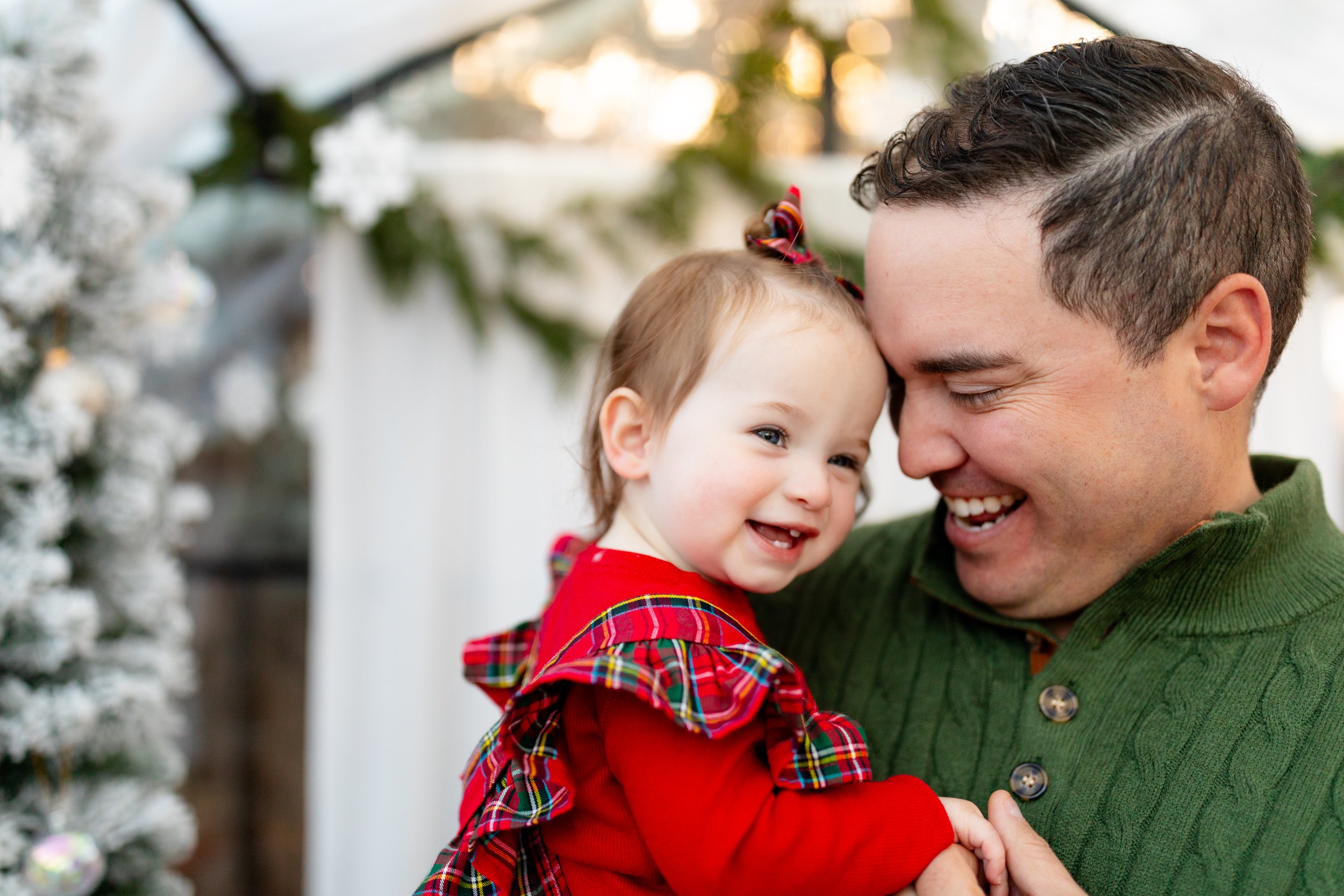 A man holding a young girl dressed in a red festive outfit, both smiling and touching foreheads, with Christmas decorations in the background.