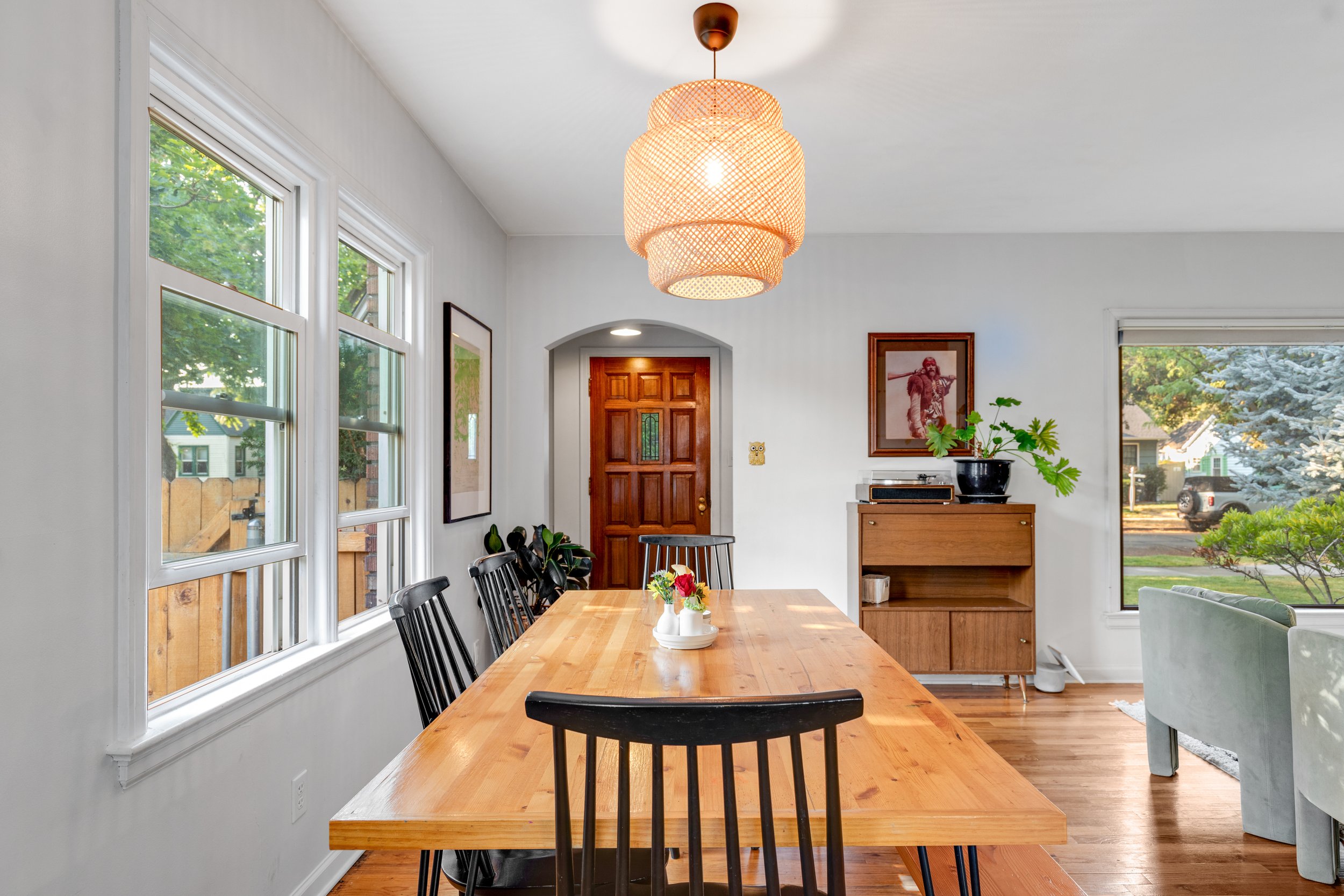 Dining room with a wooden table surrounded by black chairs, a wooden sideboard, large windows, and a woven pendant light fixture hanging from the ceiling.