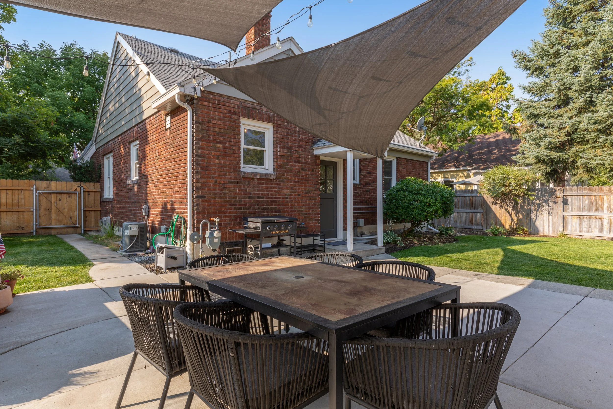 Backyard patio with wooden dining table and six black chairs, shaded by tan sun sails, adjacent to a brick house with a small porch, grill, outdoor utilities, and landscaped lawn with trees and bushes, enclosed by a wooden fence.