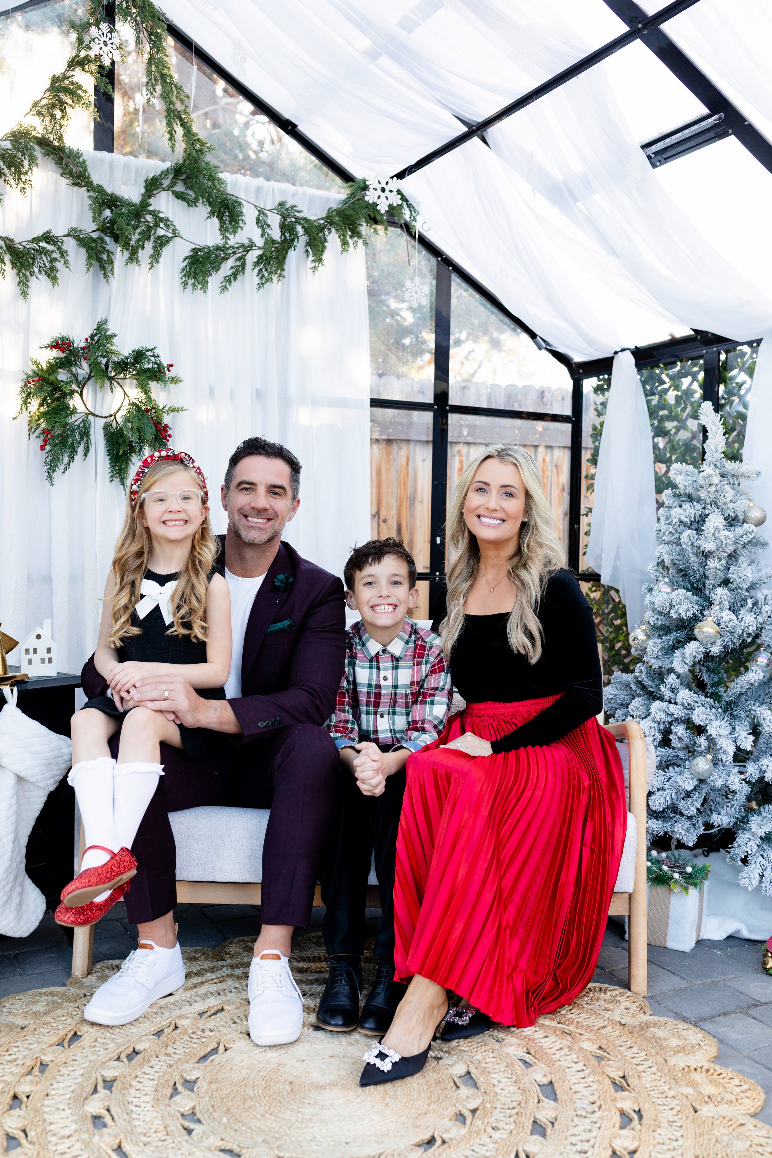 A family of four sitting together, celebrating a holiday in a decorated glass-enclosed outdoor space with Christmas decorations including a Christmas tree, a wreath, and snowflake ornaments.