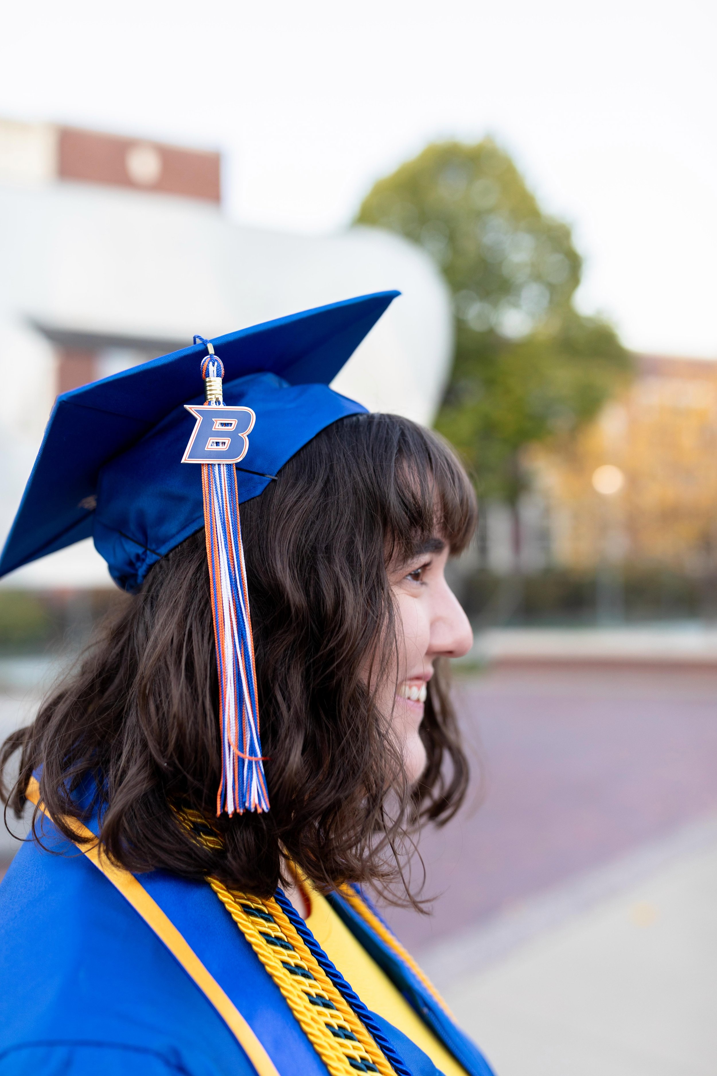 A young woman with brown hair wearing a blue graduation cap and gown, adorned with academic cords and a tassel with a 'B' charm, standing outdoors during daytime.