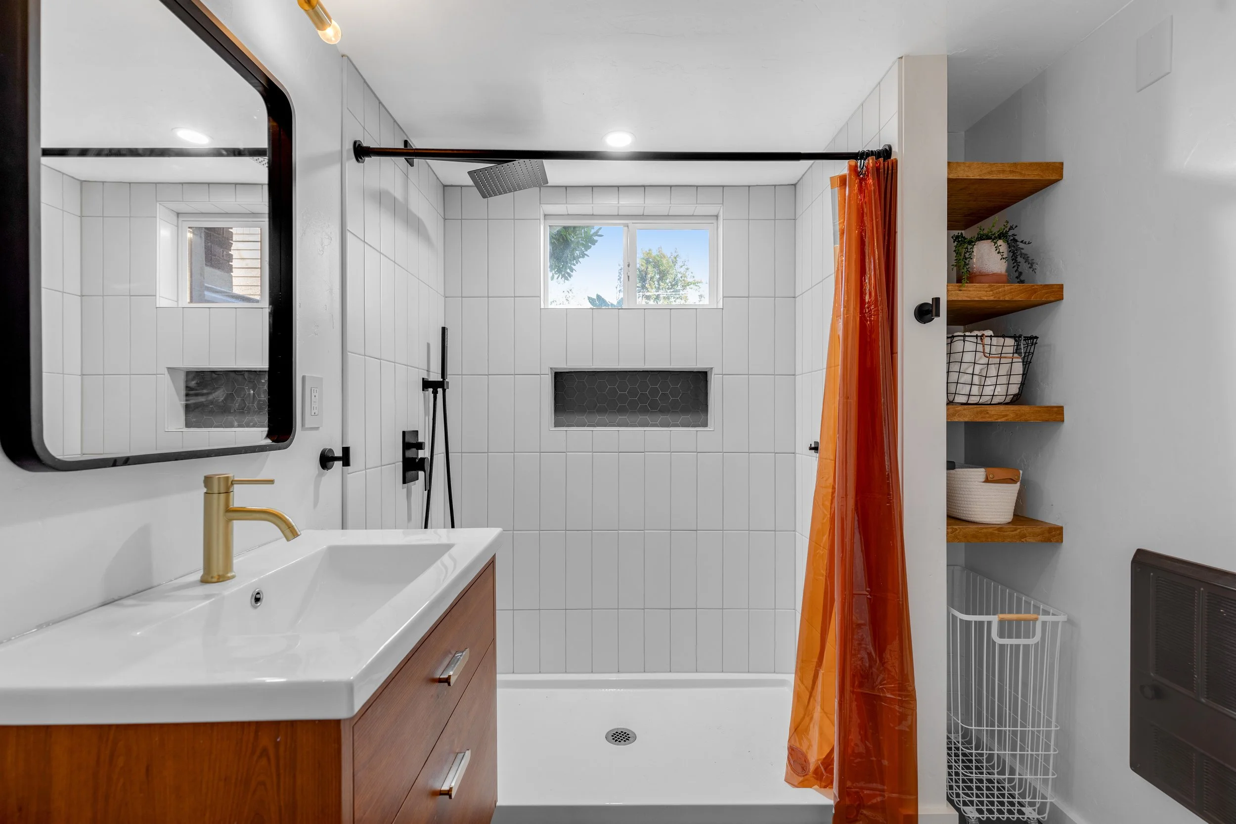 Modern bathroom with walk-in shower featuring white tiled walls, black fixtures, a small window, and a wood vanity with a white sink. An orange shower curtain is partially drawn, and wooden shelves with decor and storage baskets are on the side.