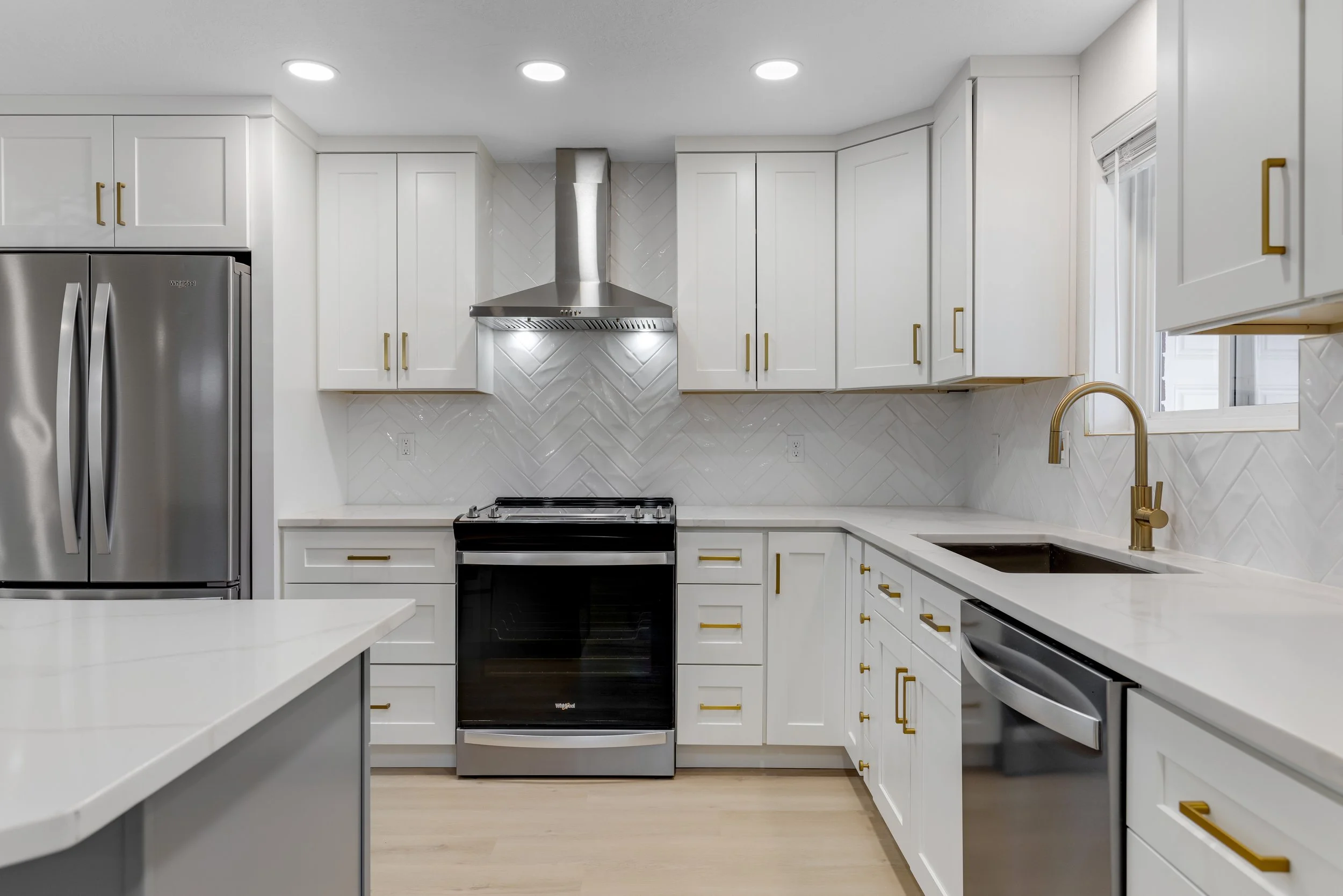 Modern white kitchen with stainless steel appliances, gold handles, backsplash, white countertops, and a gold faucet.