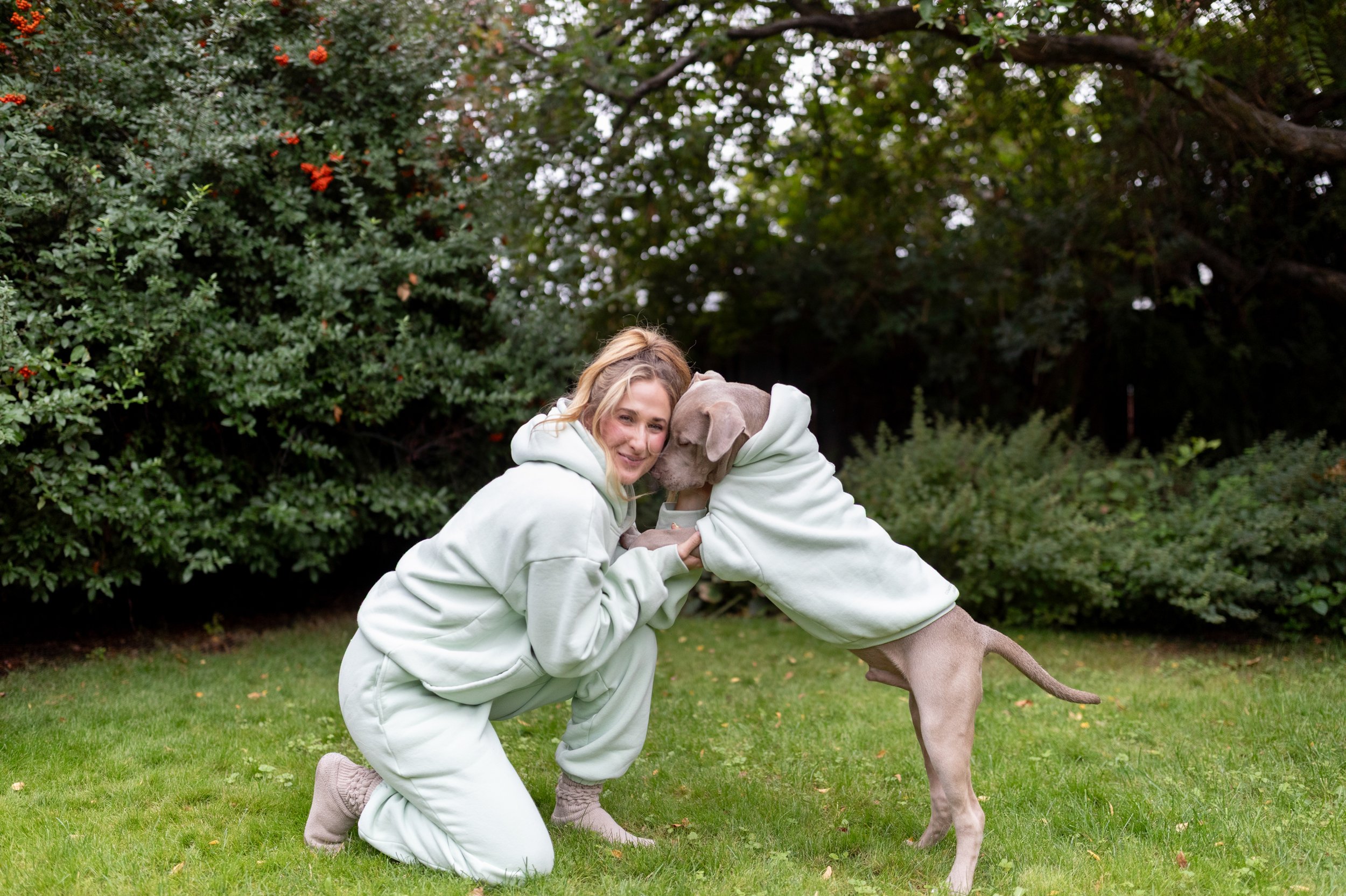 A woman and a dog both wearing matching light-colored hoodies playing together outdoors on a grassy area with bushes and trees in the background.