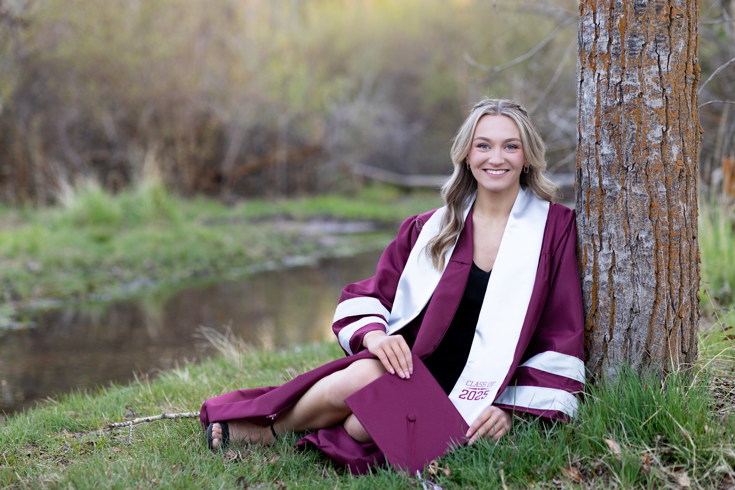 A young woman in a maroon graduation gown and cap sitting on grass beside a tree near a creek, smiling outdoors during daytime.