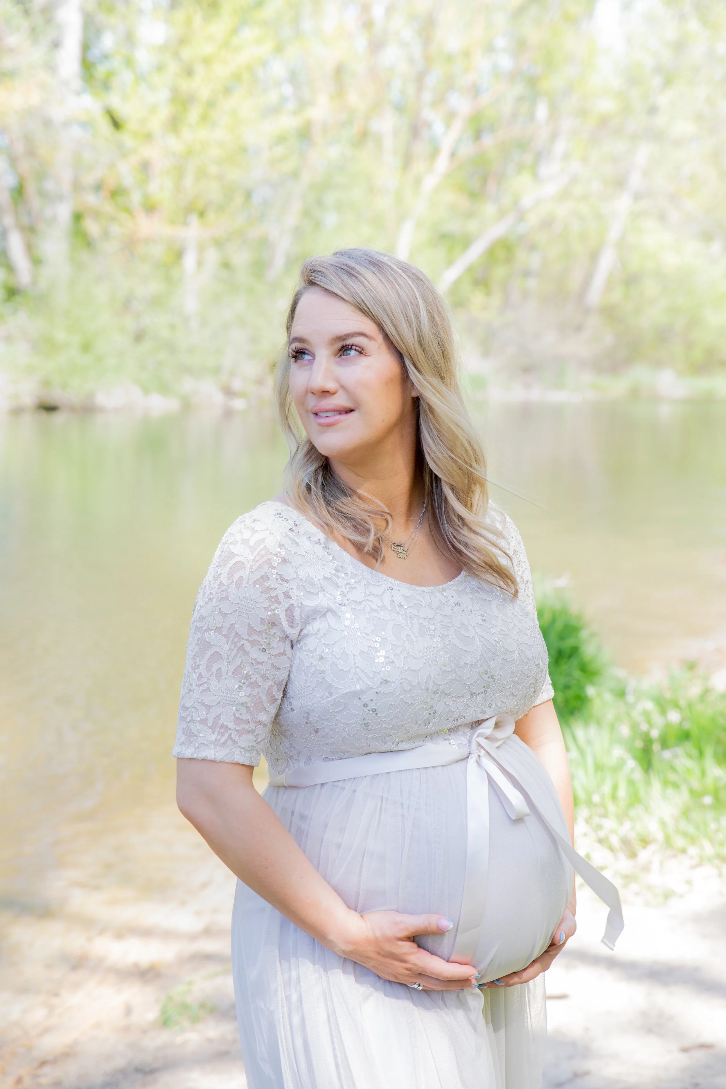Pregnant woman outdoors by a river, wearing a white lace top and smiling.