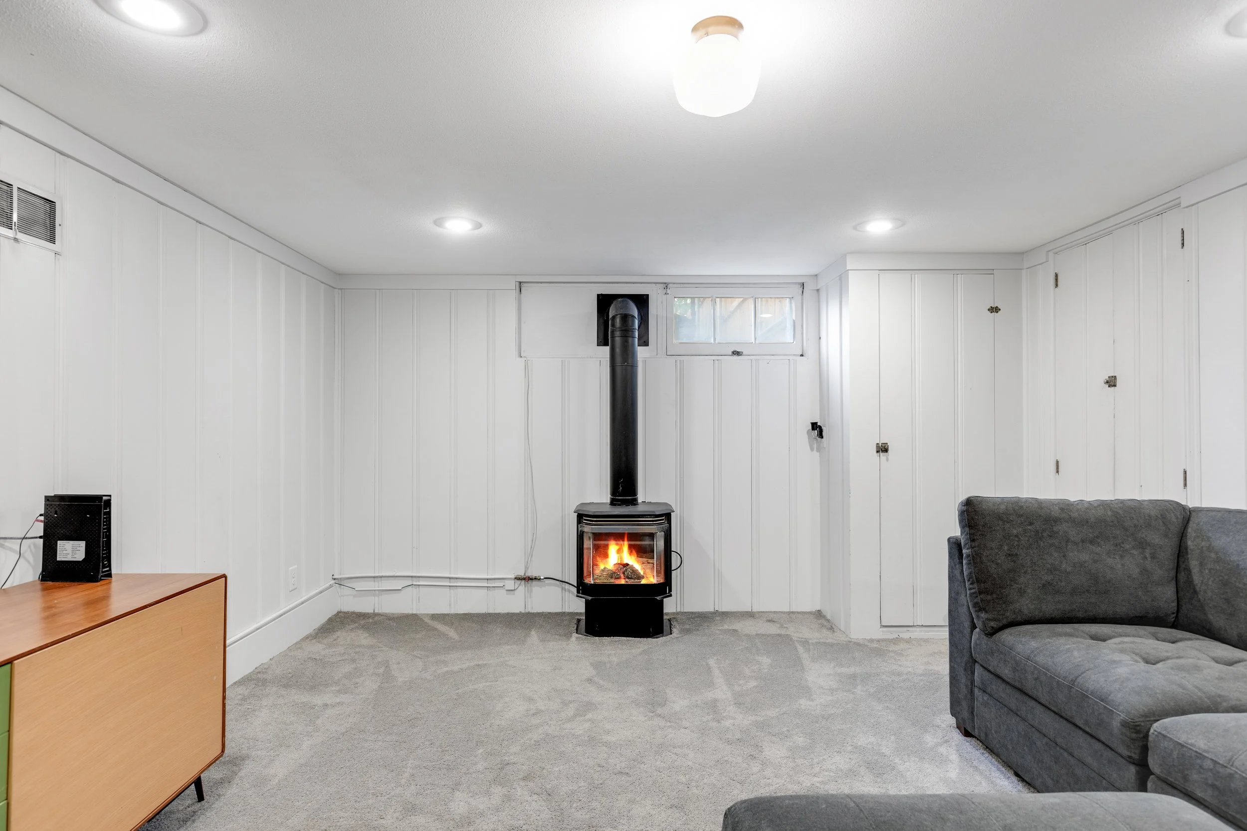 Living room with gray sofa, wood cabinet, wall heater, and a black wood stove with a pipe, on a beige carpet, white paneled walls, and small window.