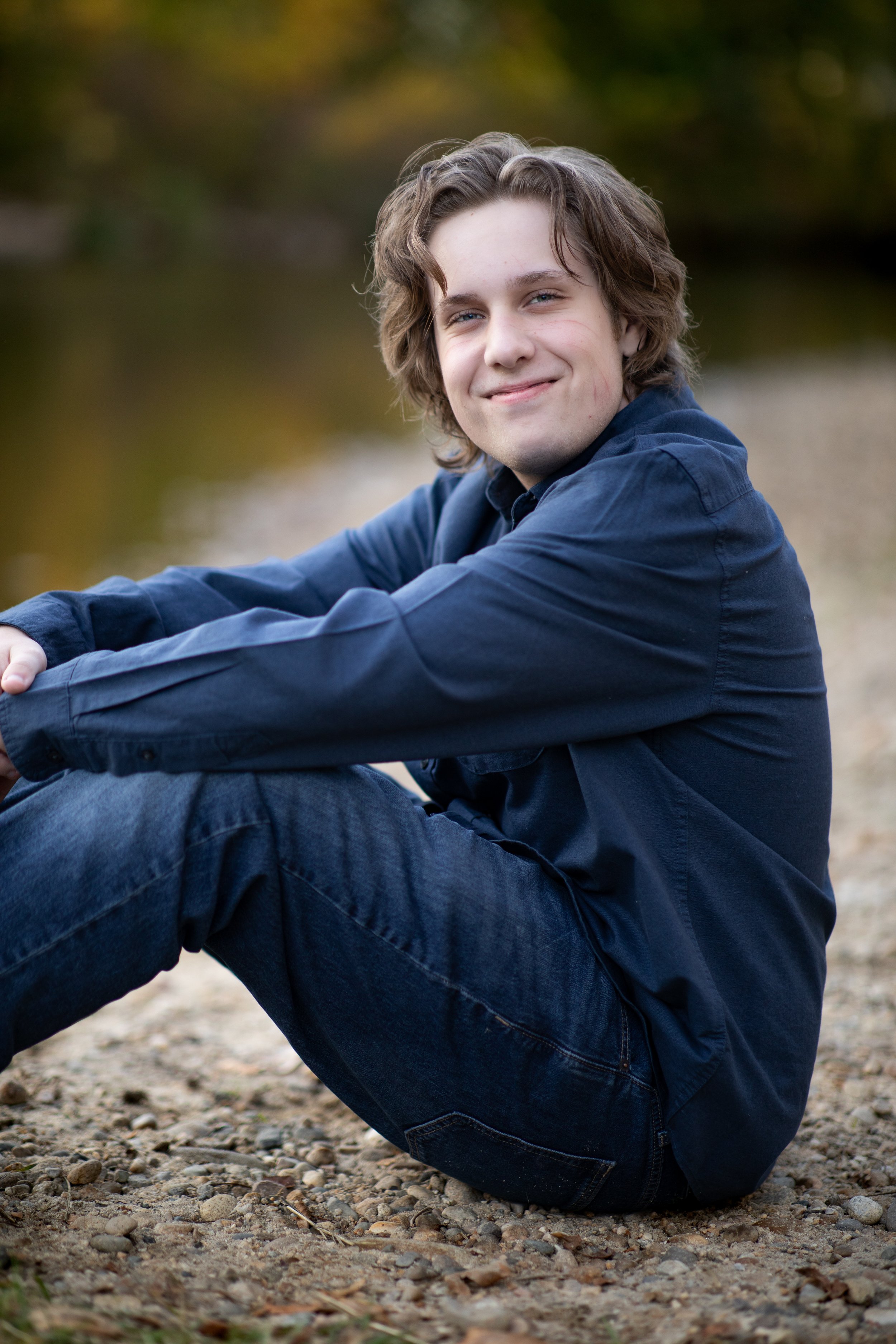 A young man with wavy brown hair and light skin sitting on a rocky shore, smiling at the camera with a body of water and blurred trees in the background.