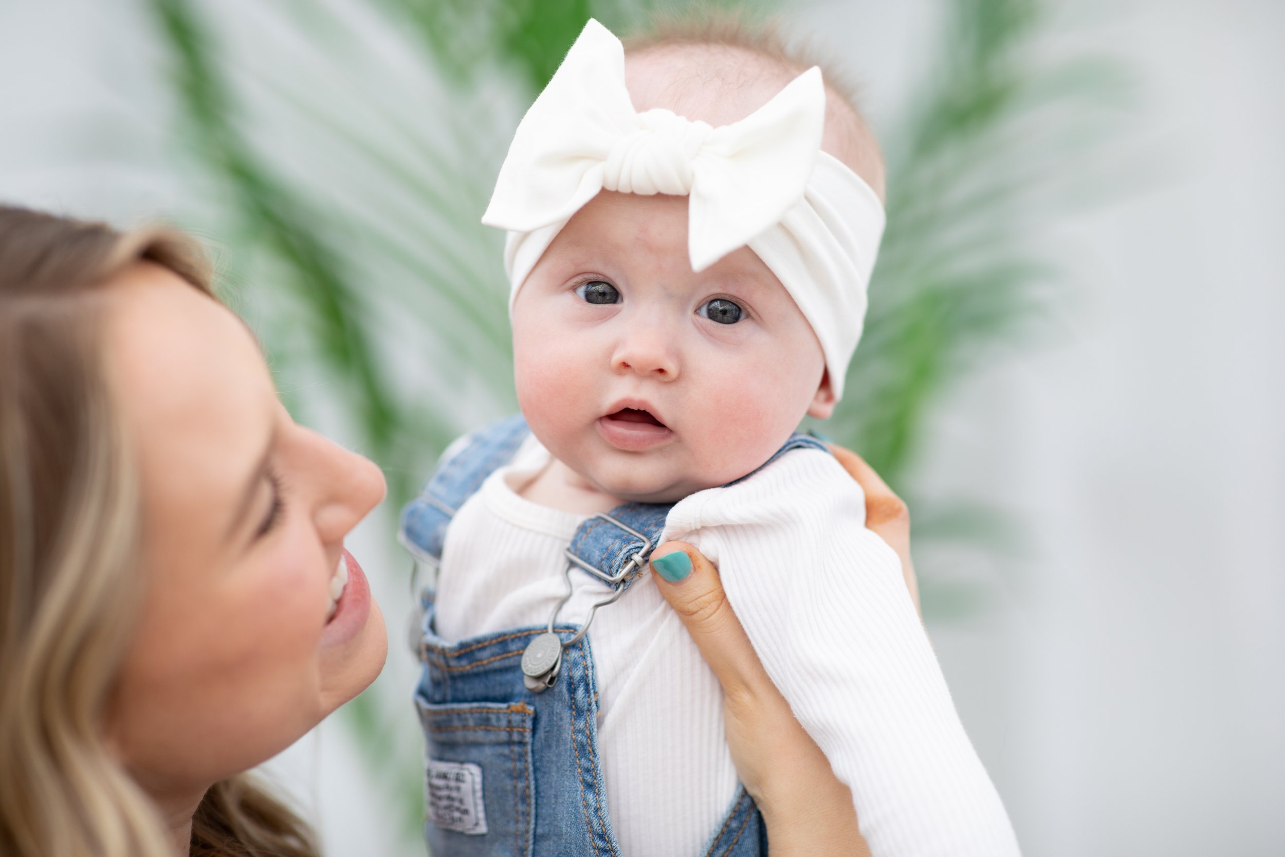 Young woman holding a baby girl with a white headband and denim overalls, smiling at her against a green background.