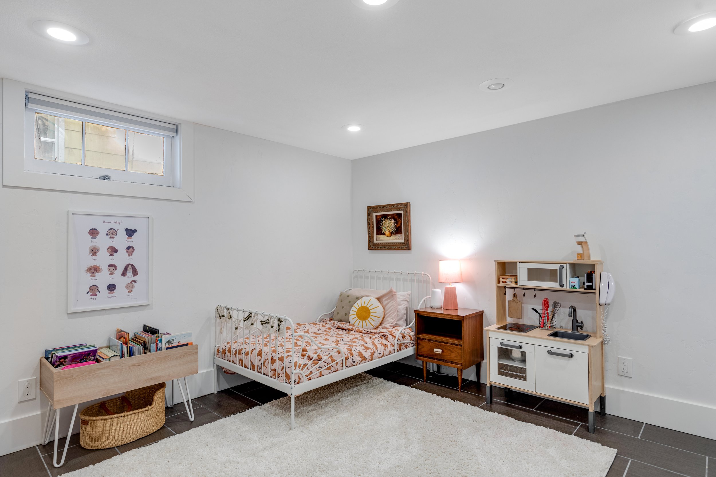 A bedroom with white walls and ceiling, a small window, a bed with beige bedding and sun-shaped pillow, a wooden nightstand with a pink lamp, a white toy kitchen set, a bookshelf with books, colorful artwork on the wall, and a white rug on a dark til