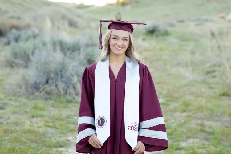 A young woman taking graduation photos in a maroon graduation gown and cap stands outdoors on a grassy path, smiling at the camera. She wears a white stole with maroon and gray accents, including a circular emblem and the text 'Class of 2023'.