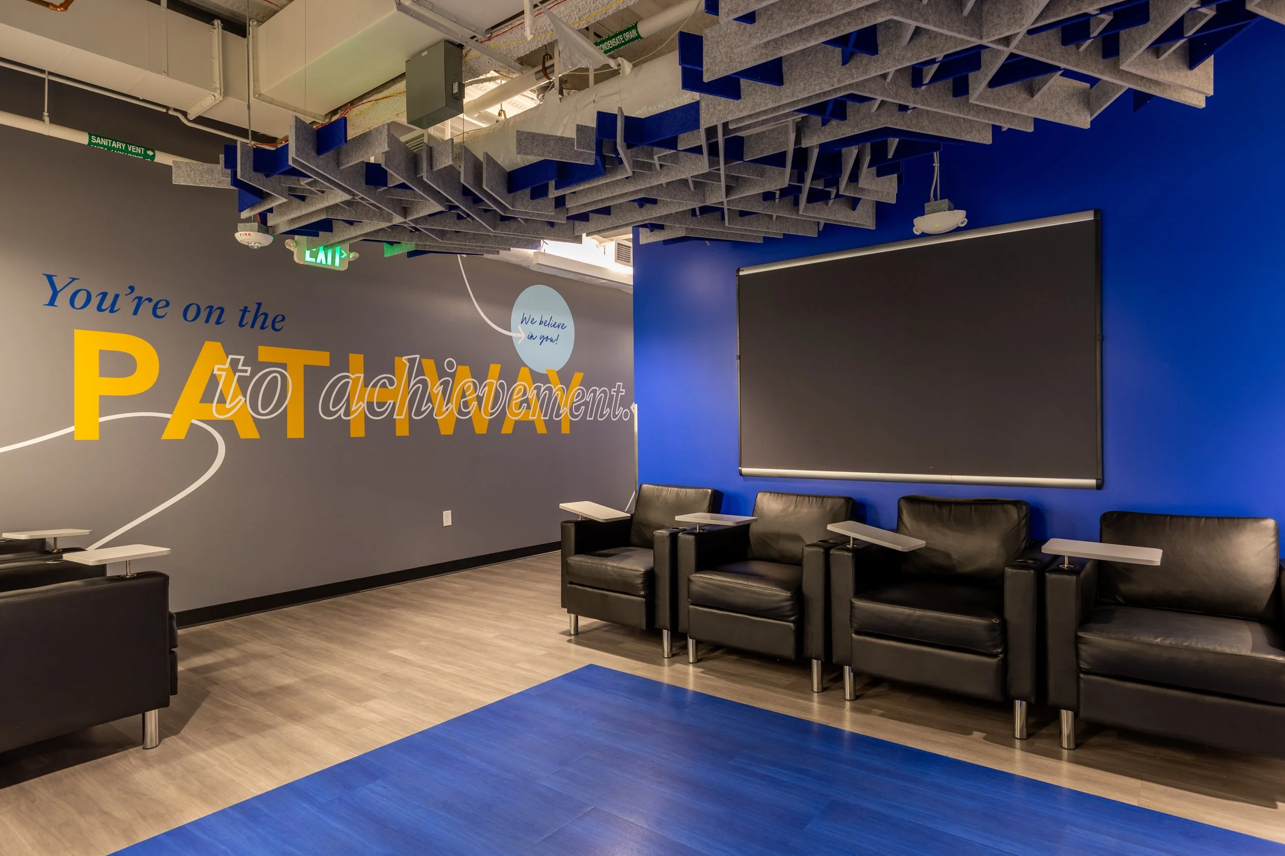 An empty waiting area with black leather chairs, a large black chalkboard, blue and gray walls, and motivational wall art that reads 'You're on the PATHWAY to achievement.'