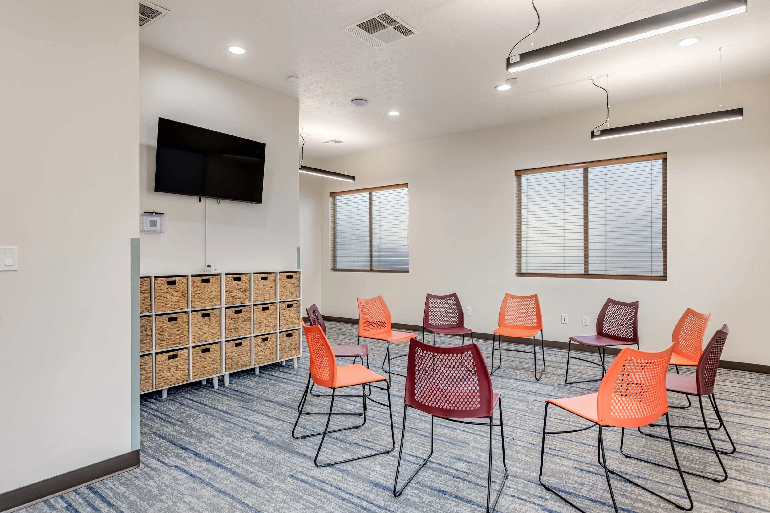 A meeting room with orange and maroon chairs arranged in a circle, a wall-mounted TV, wooden storage cubbies, windows with blinds, and ceiling lights.