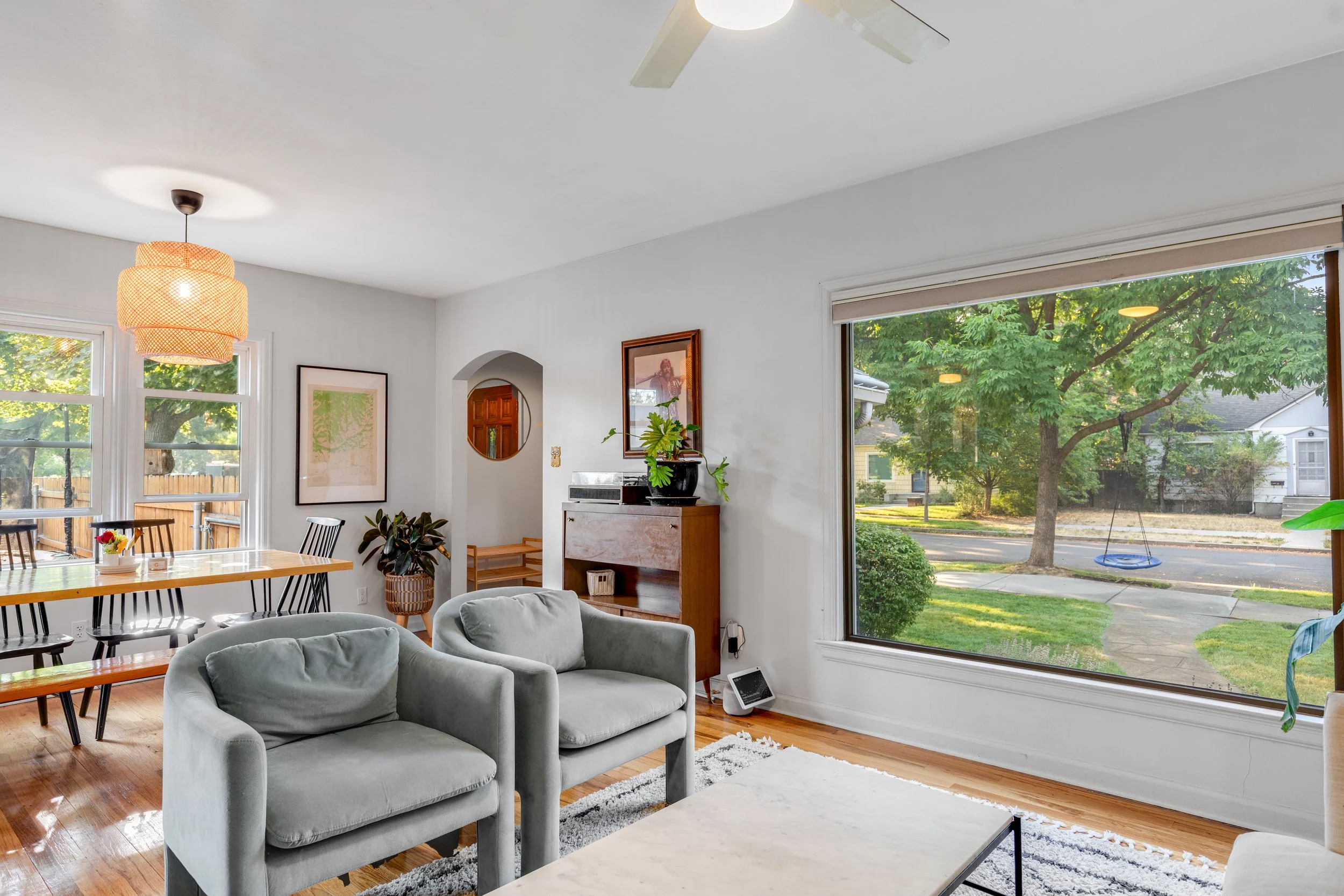 Bright living room with large front window showing an outside yard with trees and a swing, white walls, hardwood floor, light gray armchairs, dining area with wooden table and black chairs, ceiling fan, and houseplants.