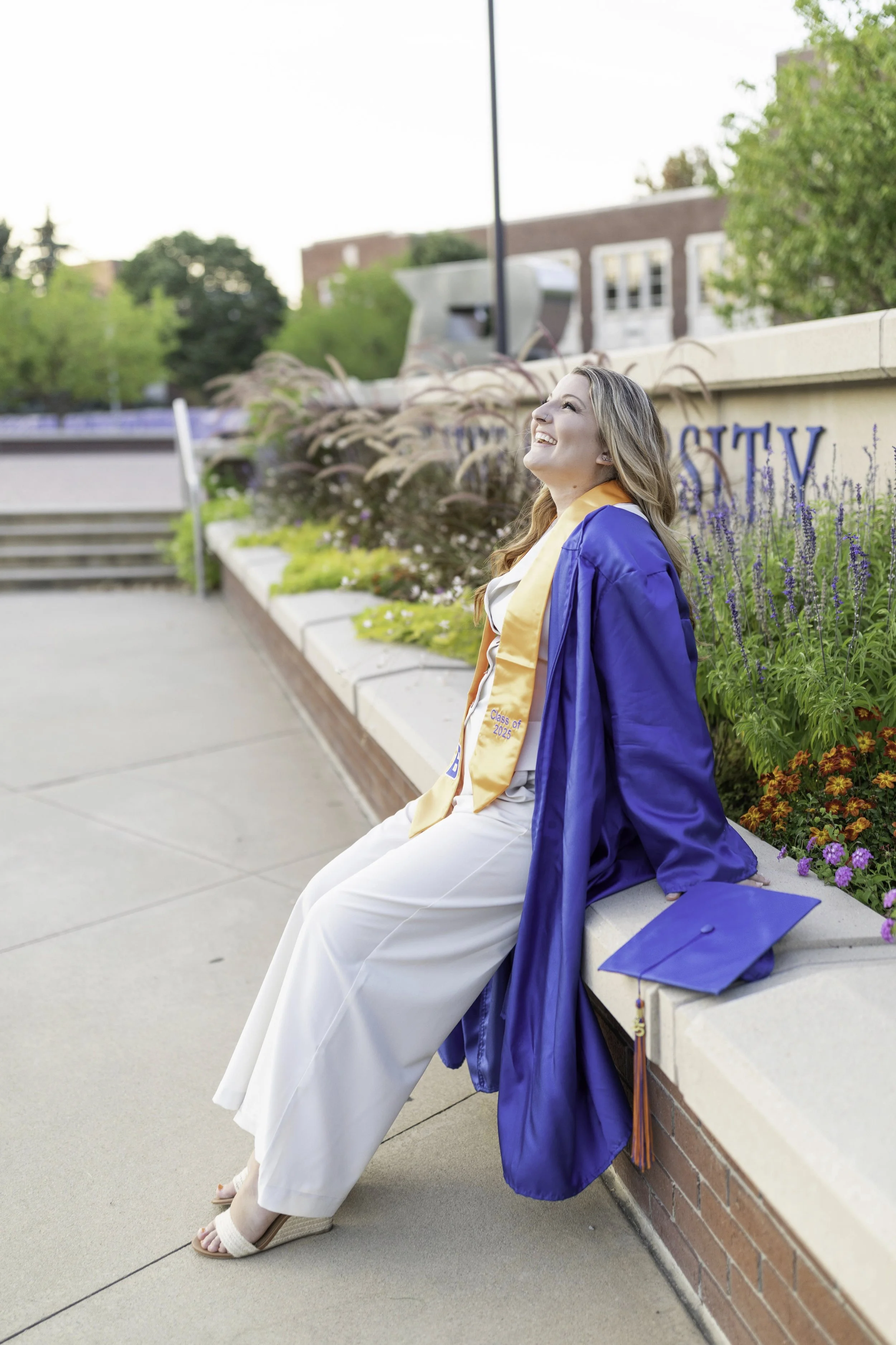 Happy woman in graduation cap and gown sitting on a low wall with a graduation stole, smiling and looking up outdoors during the daytime.