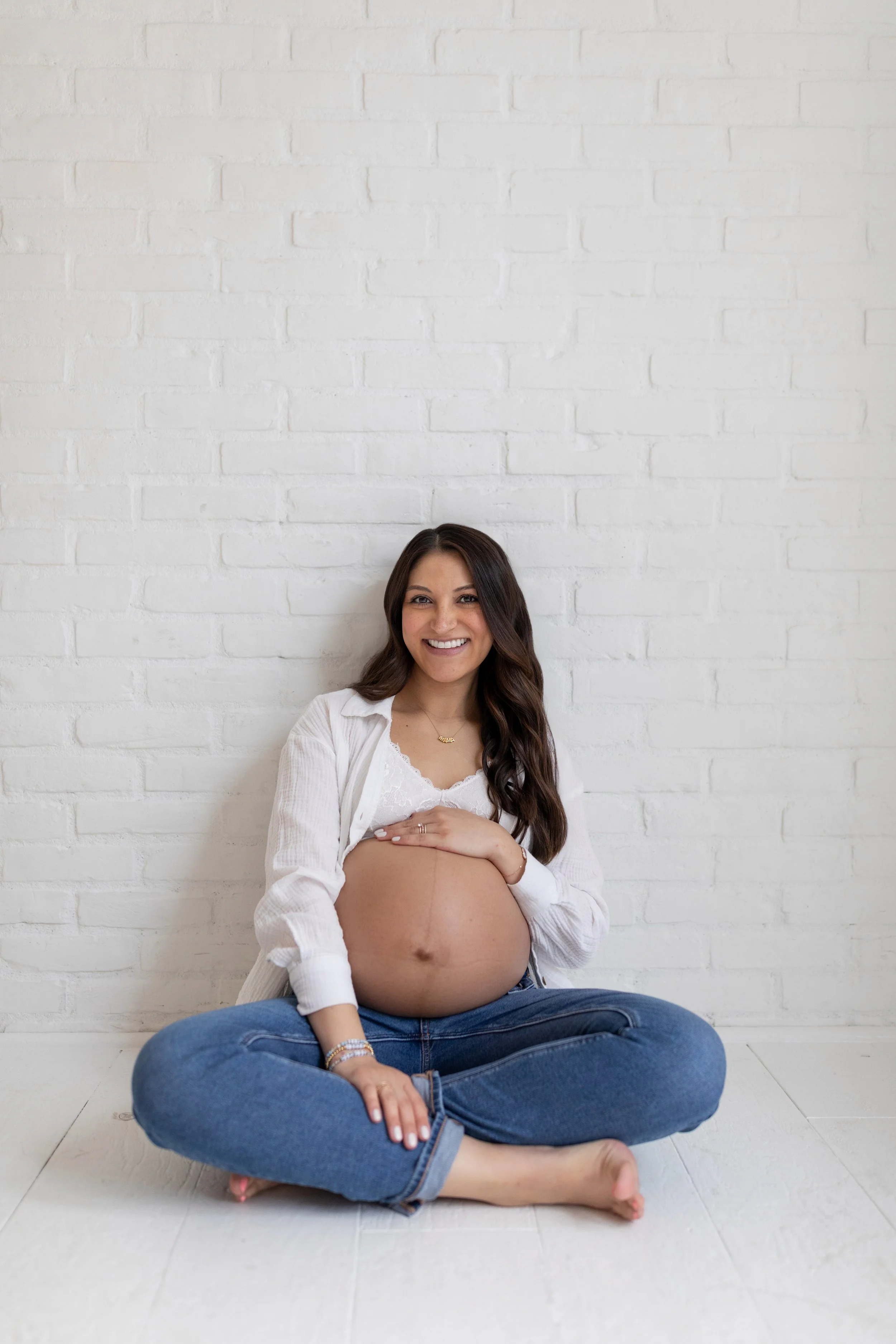 A pregnant woman sitting cross-legged against a white brick wall, smiling, wearing a white top and blue jeans.