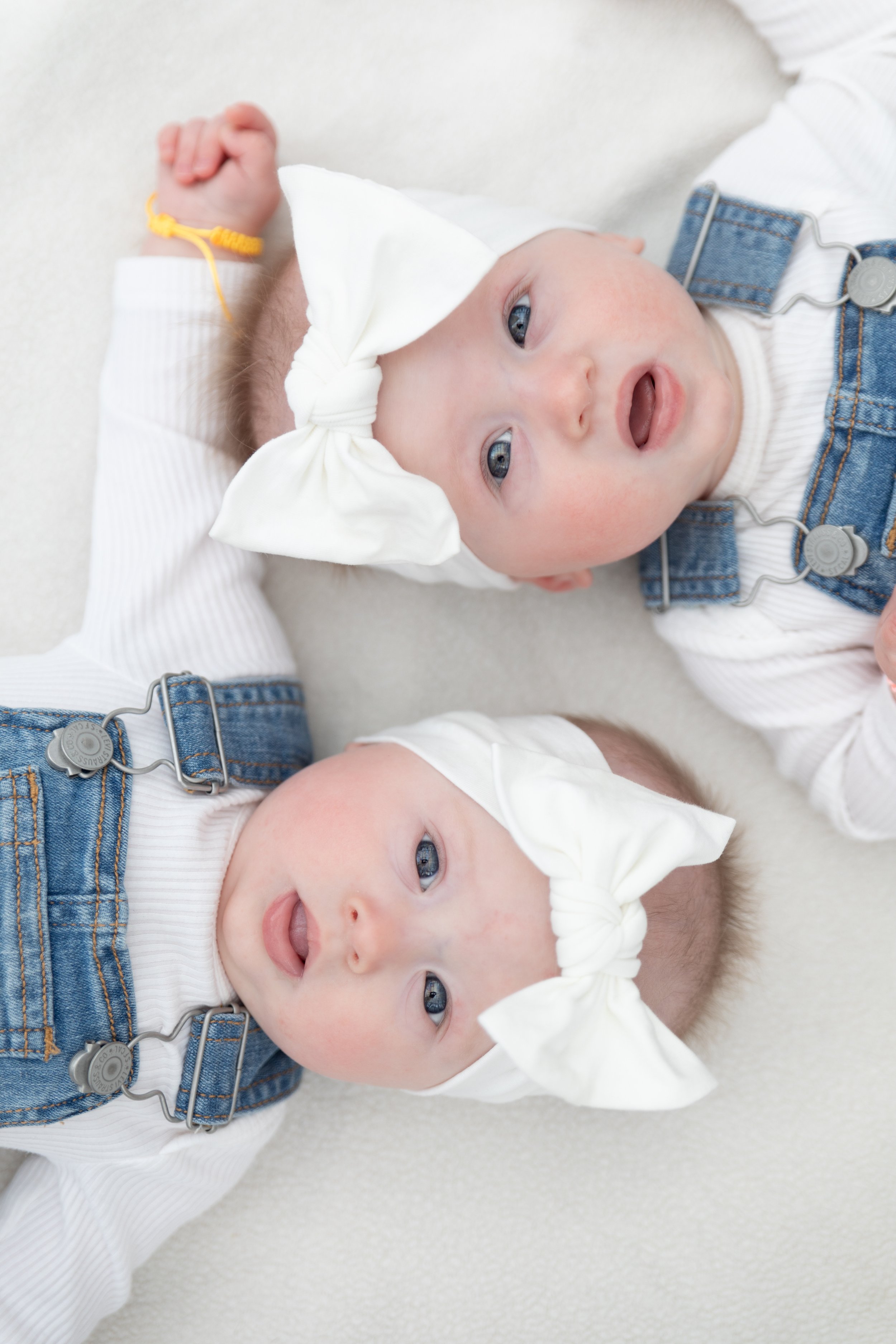 Two babies laying on their backs on a white surface, wearing white headbands with bows and denim overalls, looking up with curious expressions.