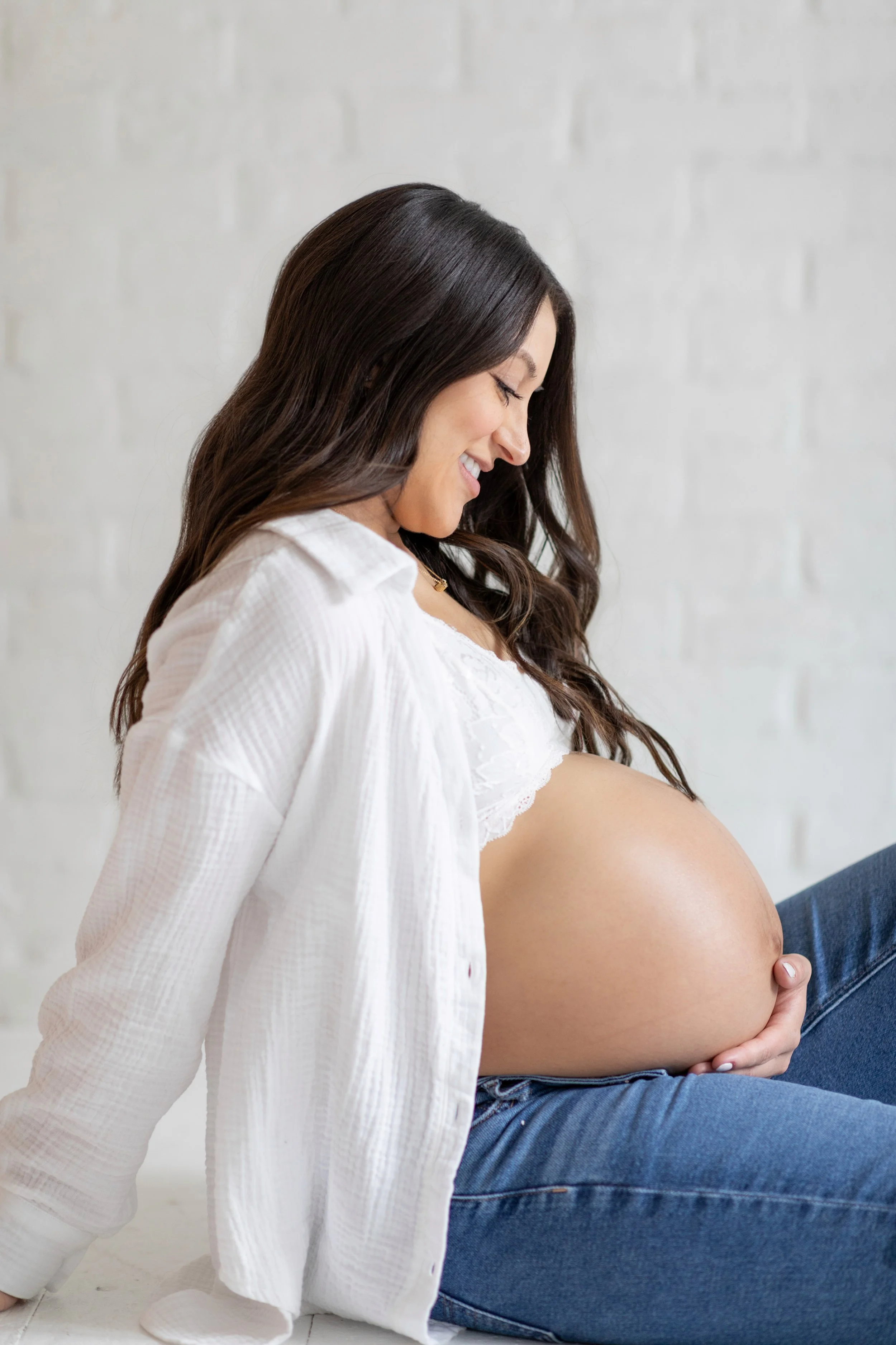 Pregnant woman smiling, sitting against a white wall, holding her belly.