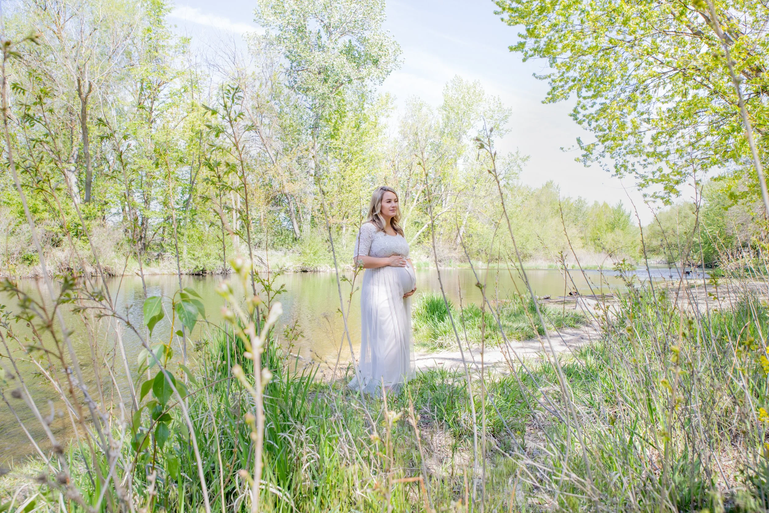 Pregnant woman standing near a river in a green forest setting on a sunny day.