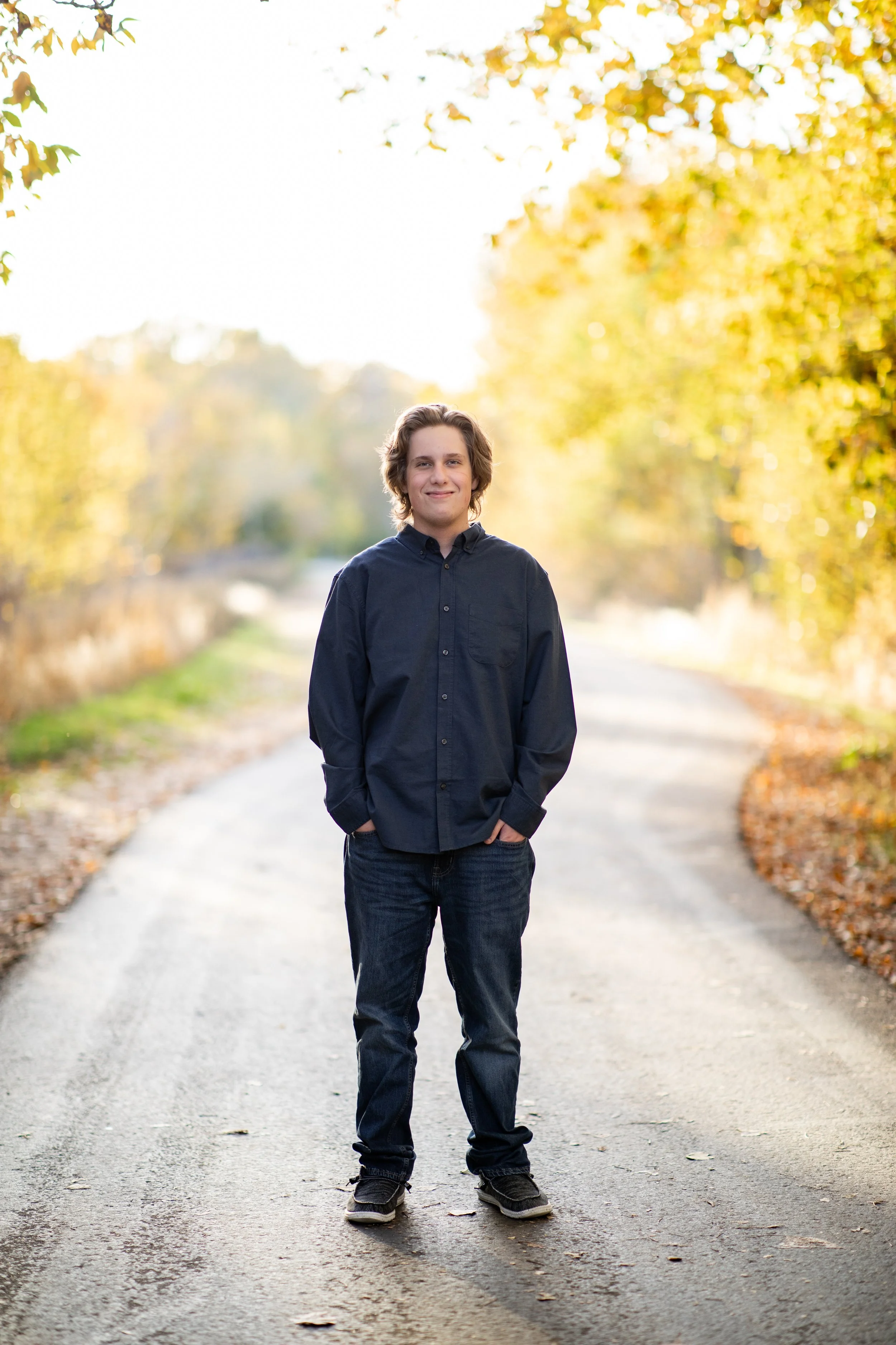 A young man with brown hair wearing a navy blue button-up shirt and jeans, standing with his hands in his pockets on a paved path surrounded by autumn-colored trees, smiling at the camera.