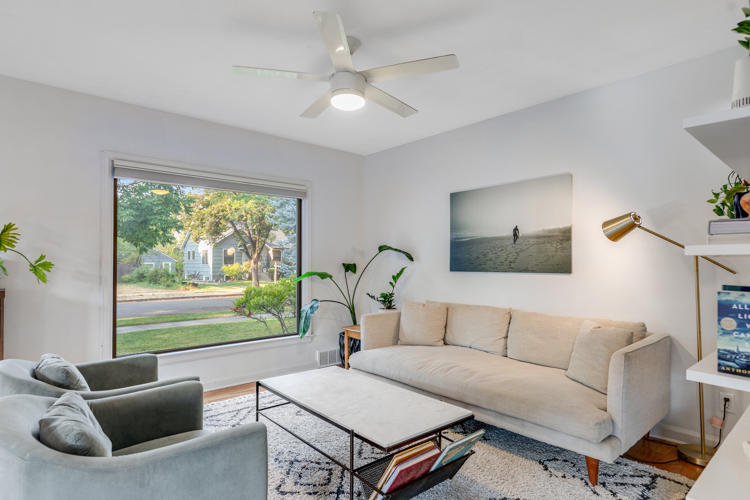 Bright living room with large window, beige sofa, green armchair, black-and-white rug, and wall art of a lone figure on the beach.