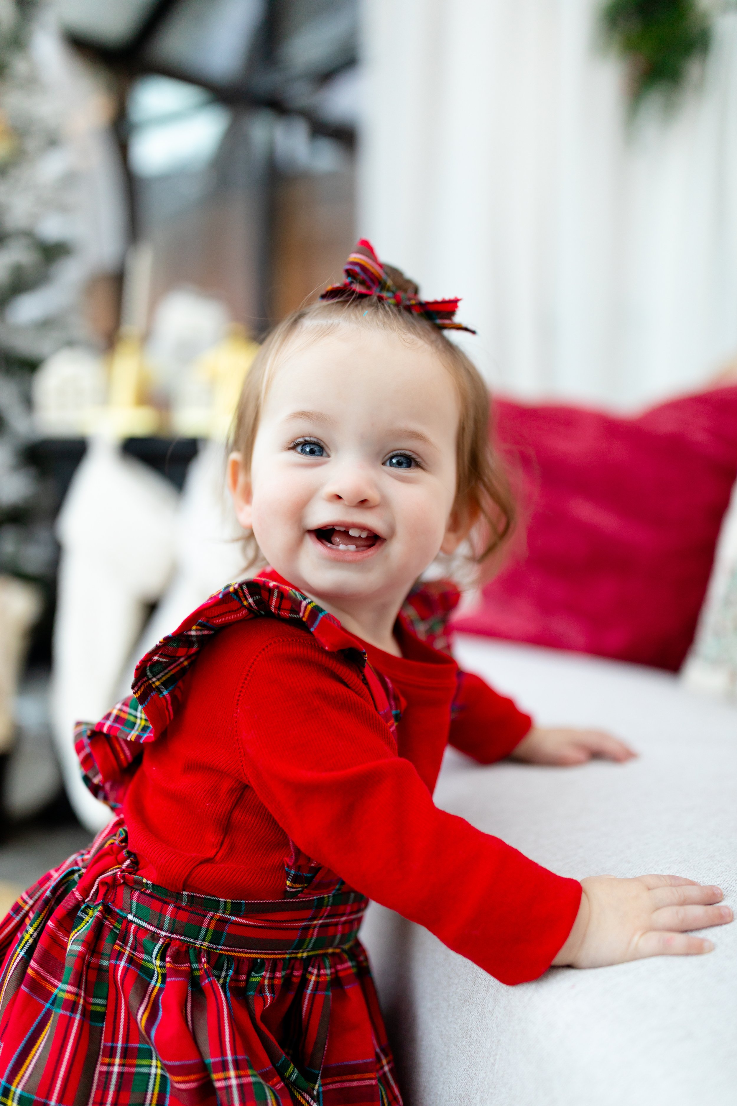 A young girl in a red dress with tartan accents, smiling and crawling on a light-colored surface, with Christmas decorations in the background.