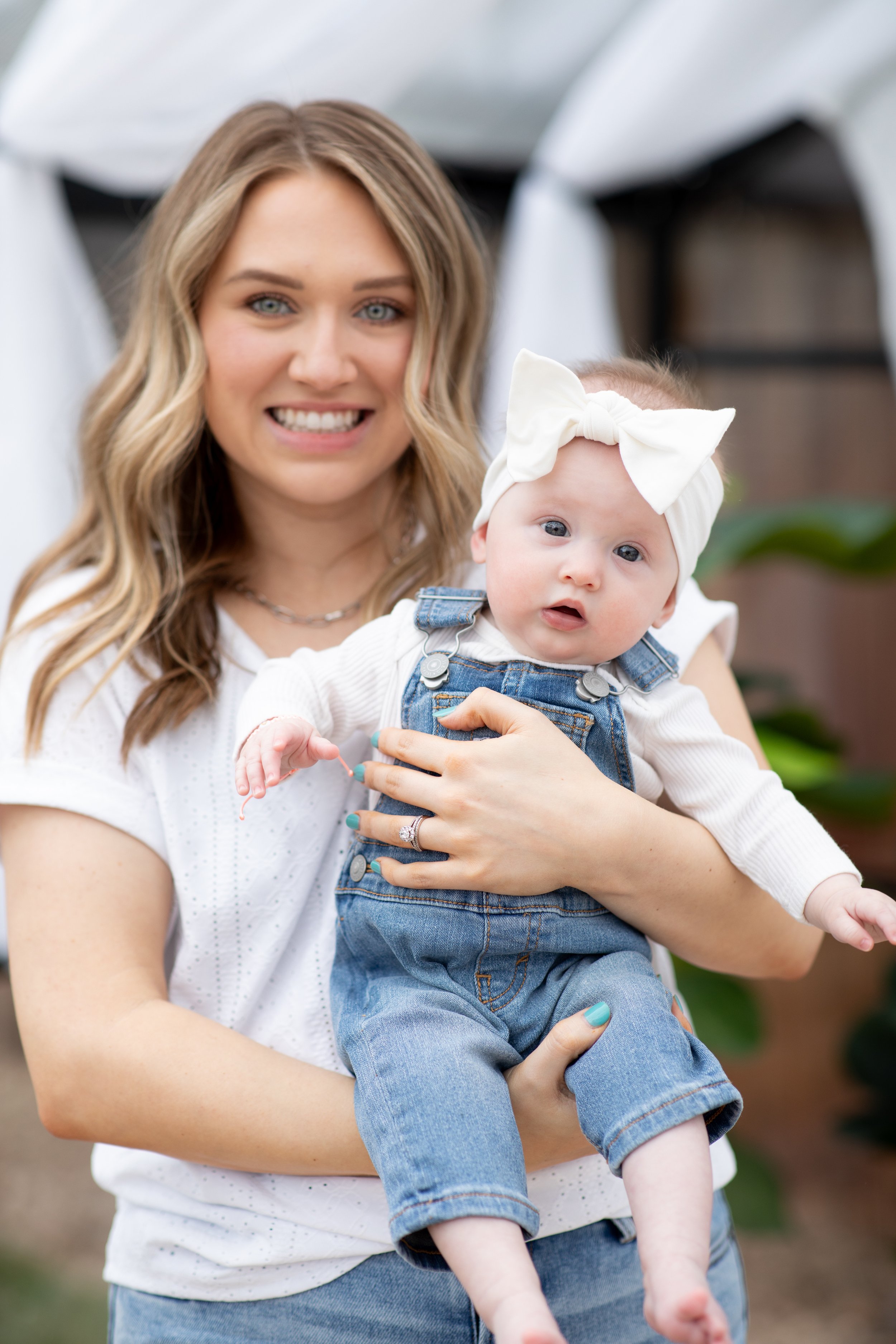 A woman with long, wavy blonde hair smiling and holding a baby girl. The baby, wearing a white headband with a bow and denim overalls, looks curiously at the camera. They are outdoors with a blurred background and a white canopy.