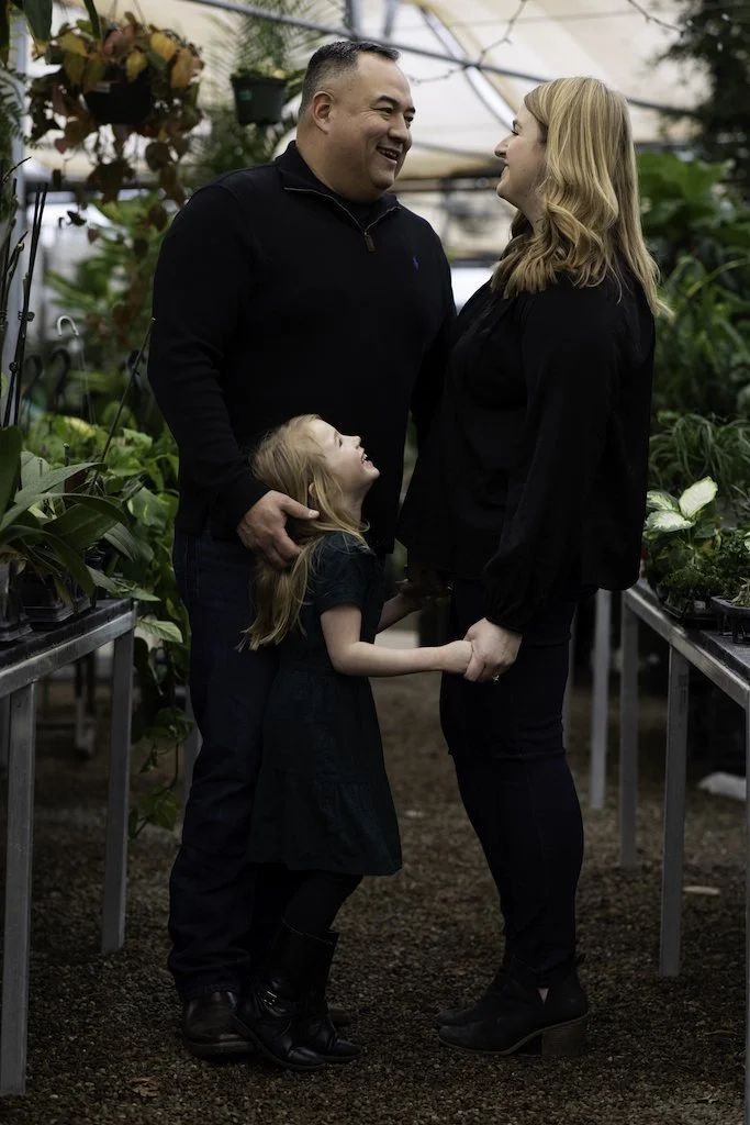 A family of three, consisting of a man, woman, and young girl, holding hands and smiling at each other inside a greenhouse filled with green plants.