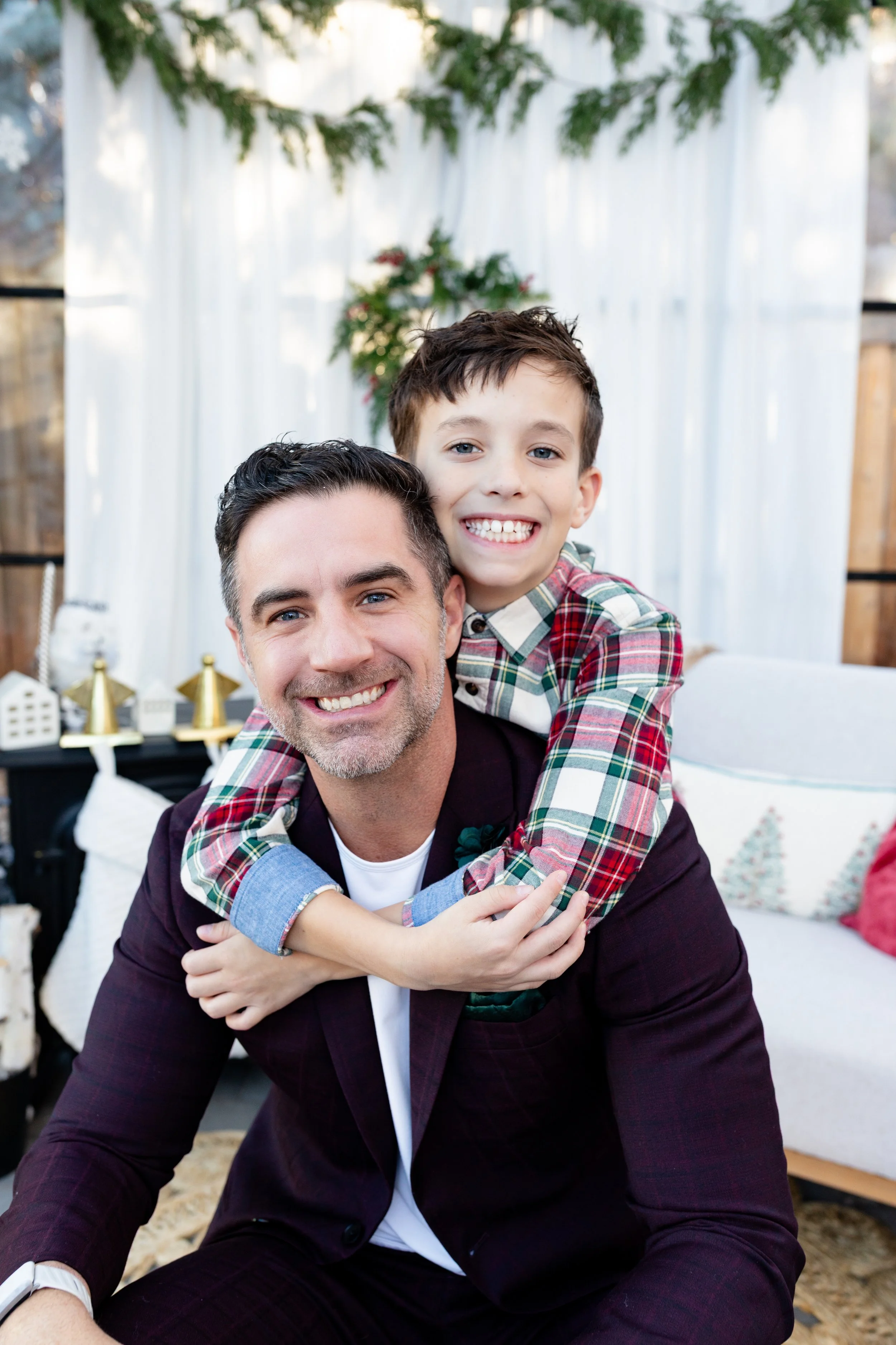 A man and a boy smiling, with the boy on his back in a piggyback ride, in a decorated indoor setting with Christmas holiday decorations, including white curtains, greenery, and small holiday-themed items.