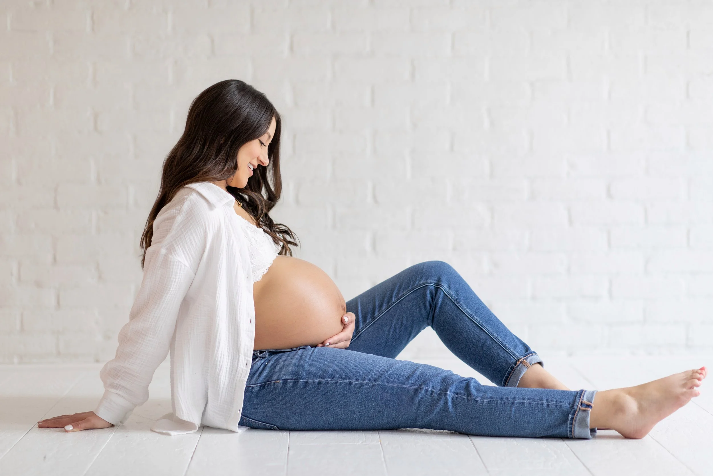 Pregnant woman sitting on the floor, smiling, holding her belly, with a white brick wall background.