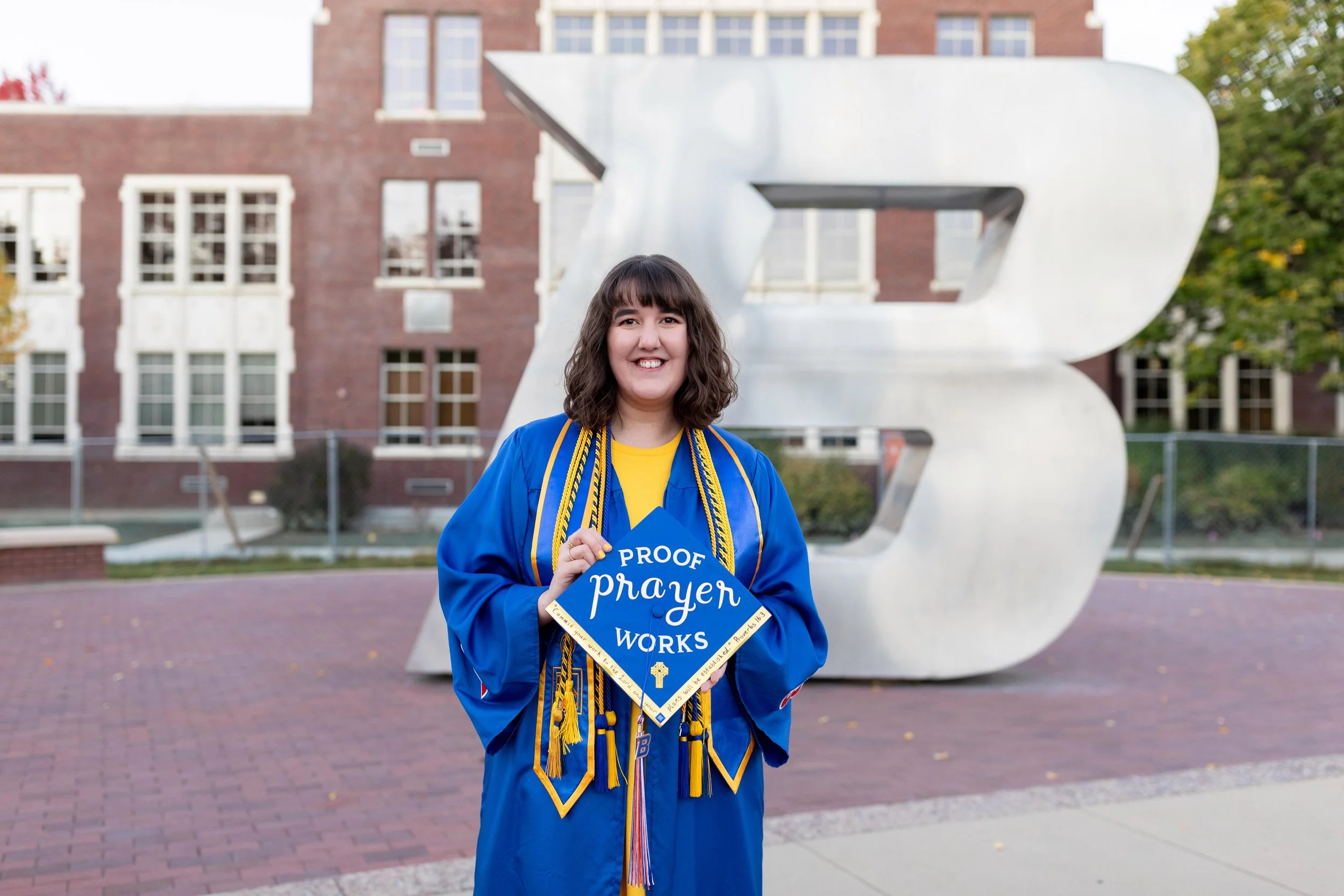 A young woman in a blue graduation gown and tassel holds a sign that reads 'Proof prayer works'. She is smiling and standing outside in front of a large, modern letter sculpture and a brick school building.
