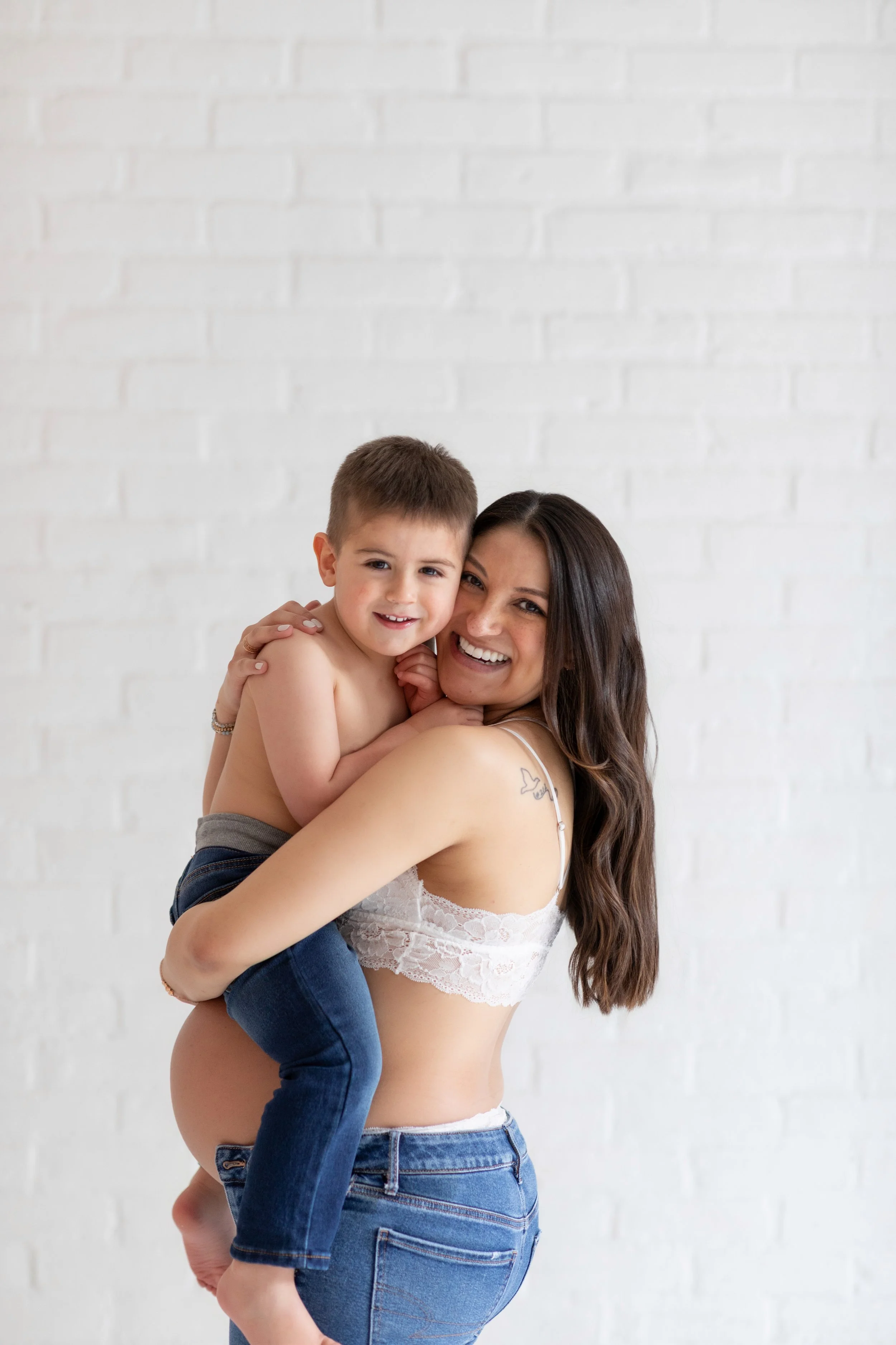 A woman with long brown hair holding a smiling young boy with short brown hair, both looking at the camera, against a white brick wall.