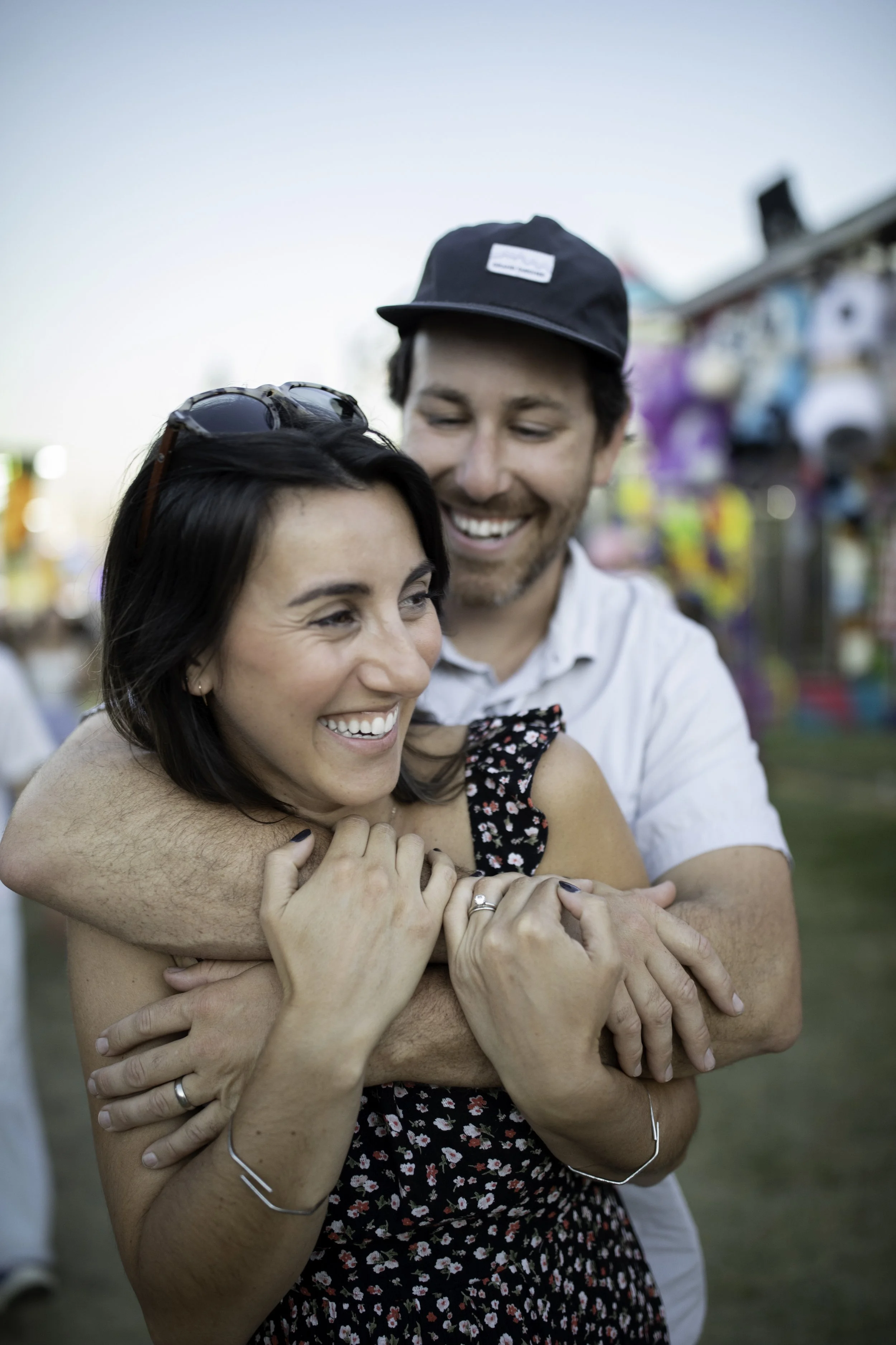 A couple hugging and smiling at a fairground or outdoor event with colorful booths and decorations in the background.