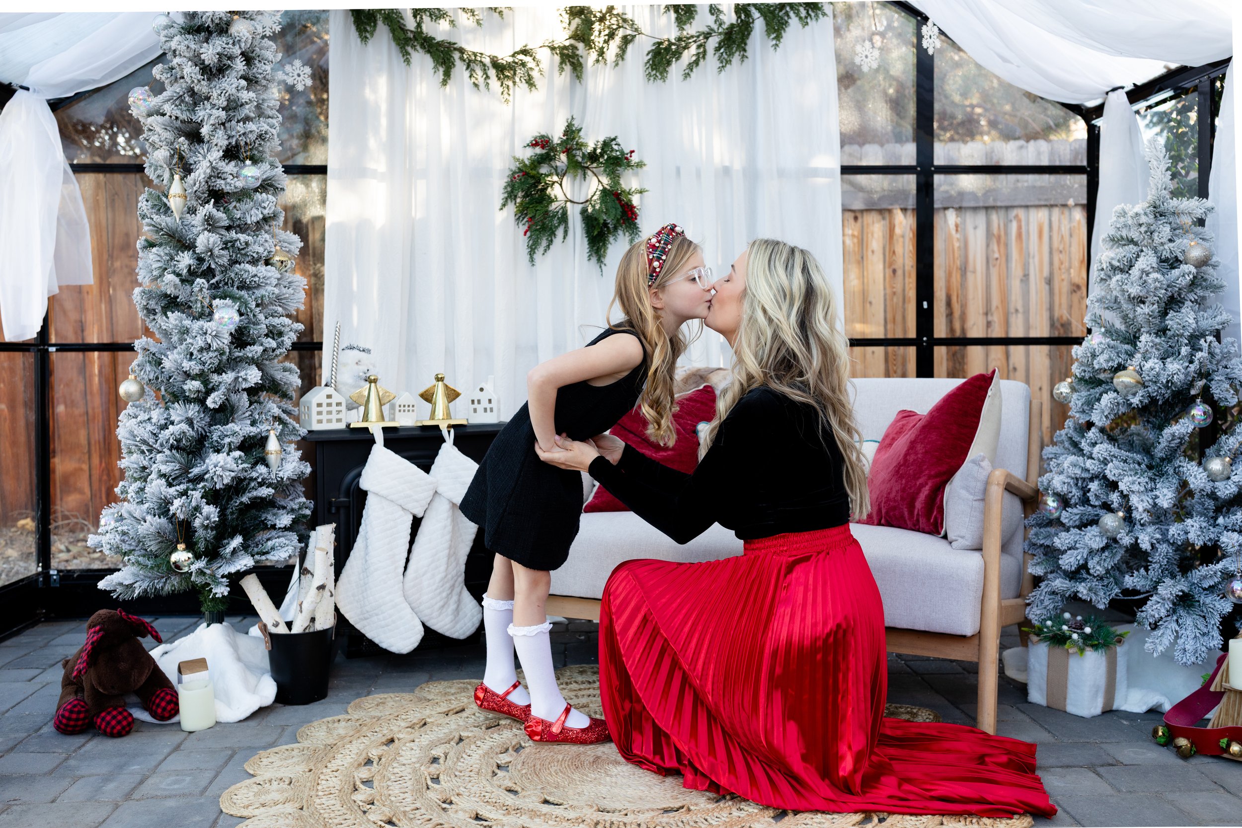 A woman kneeling and kissing a young girl, both dressed festively, in a decorated Christmas setting with snow-covered trees, stockings, and holiday decorations.