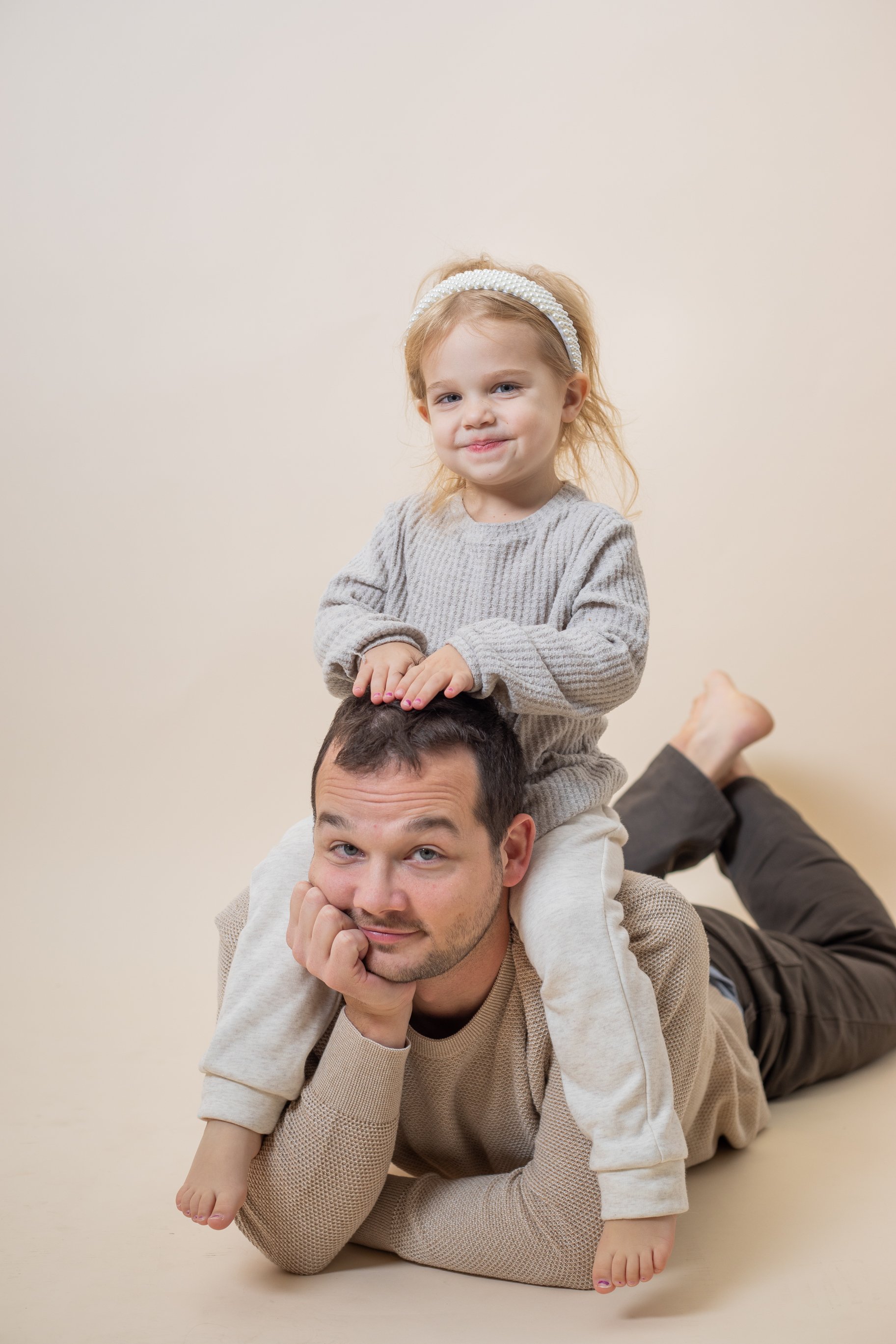 A man lying on the floor with a young girl sitting on his back, both smiling at the camera against a plain beige background.