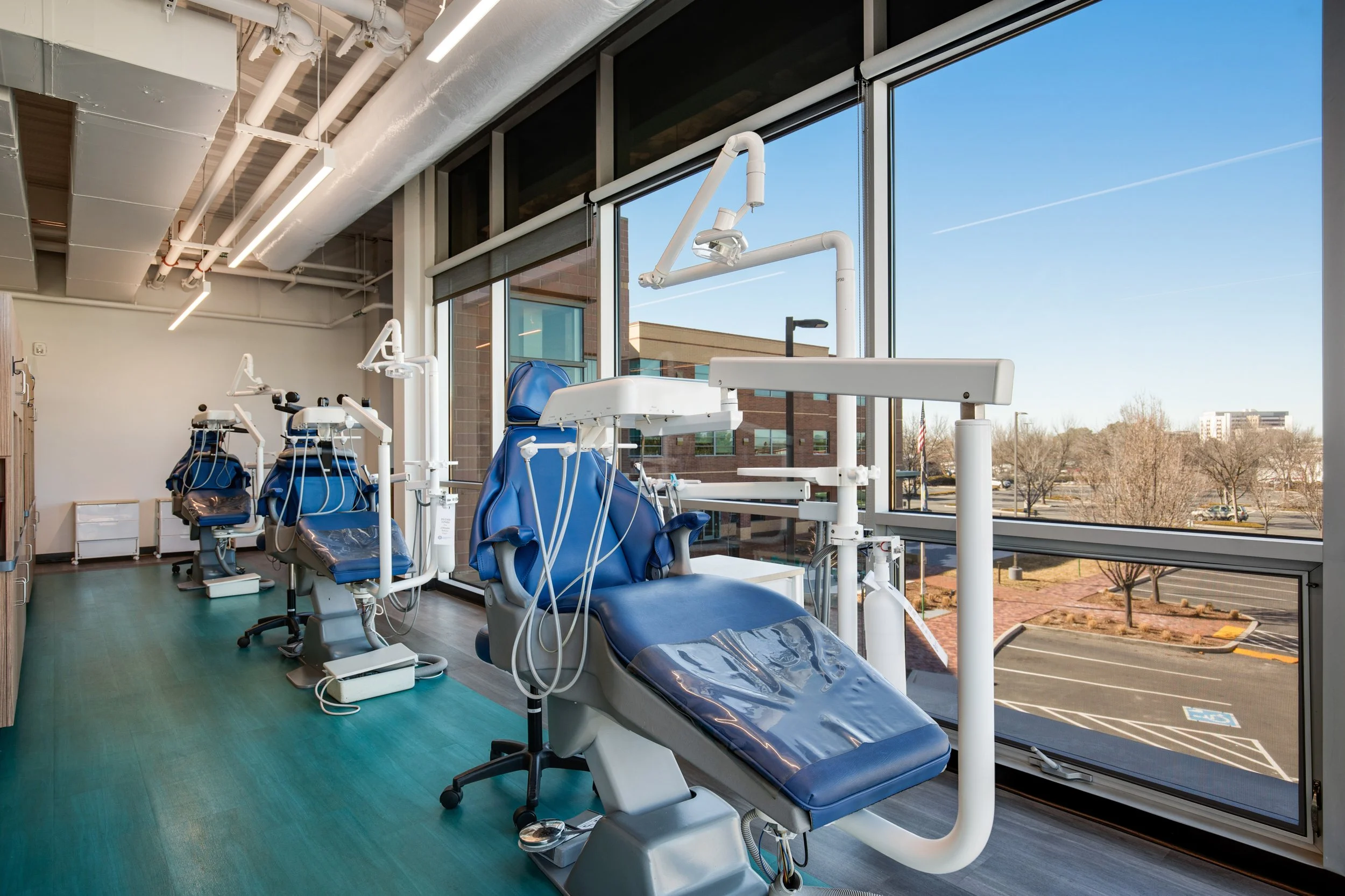 Dental treatment rooms with dental chairs near large windows overlooking a parking lot and trees, equipped with dental tools and overhead lights.