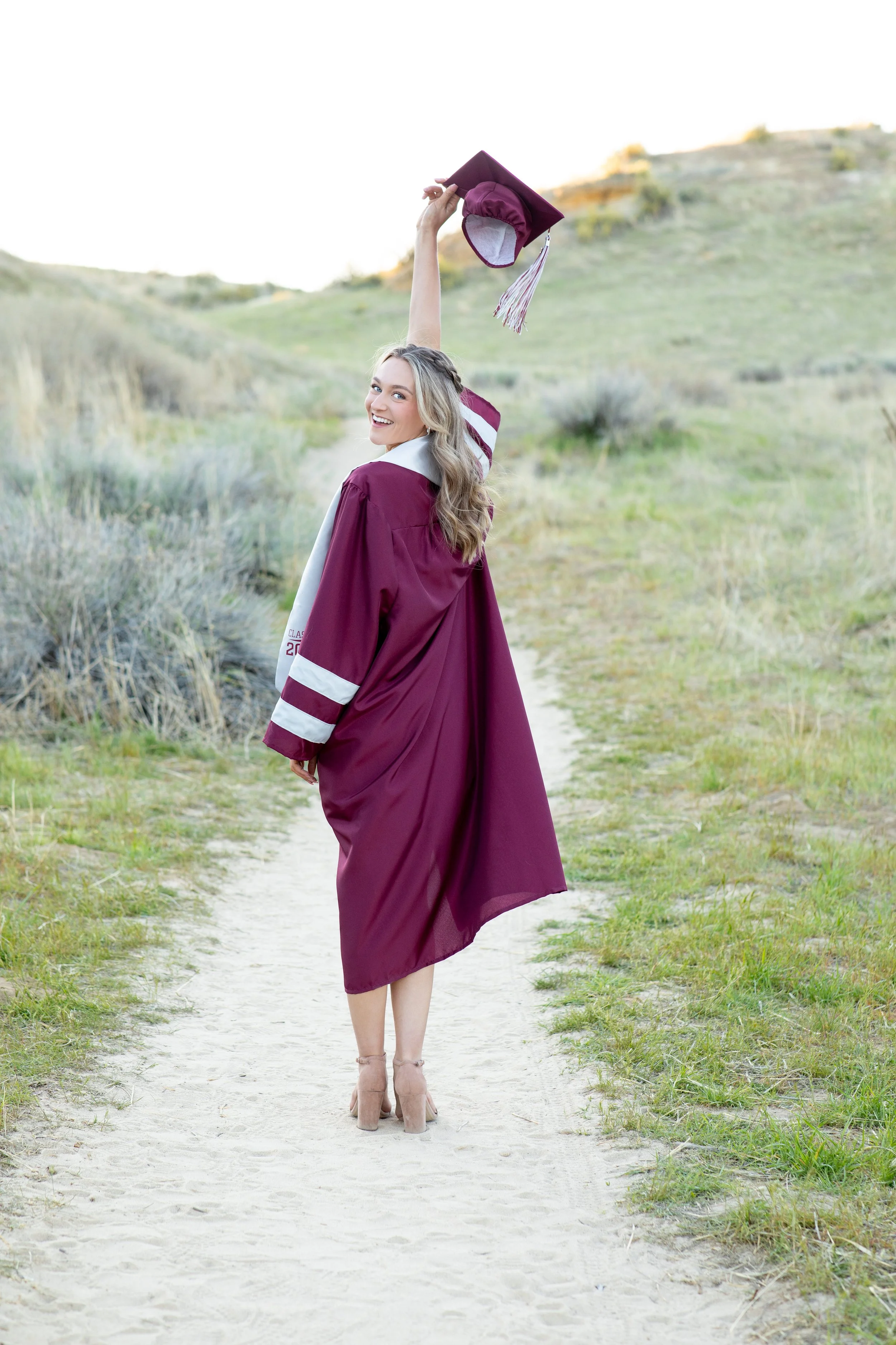 Young woman in maroon graduation gown and cap, holding cap in the air, walking along a sandy trail in a natural outdoor setting.