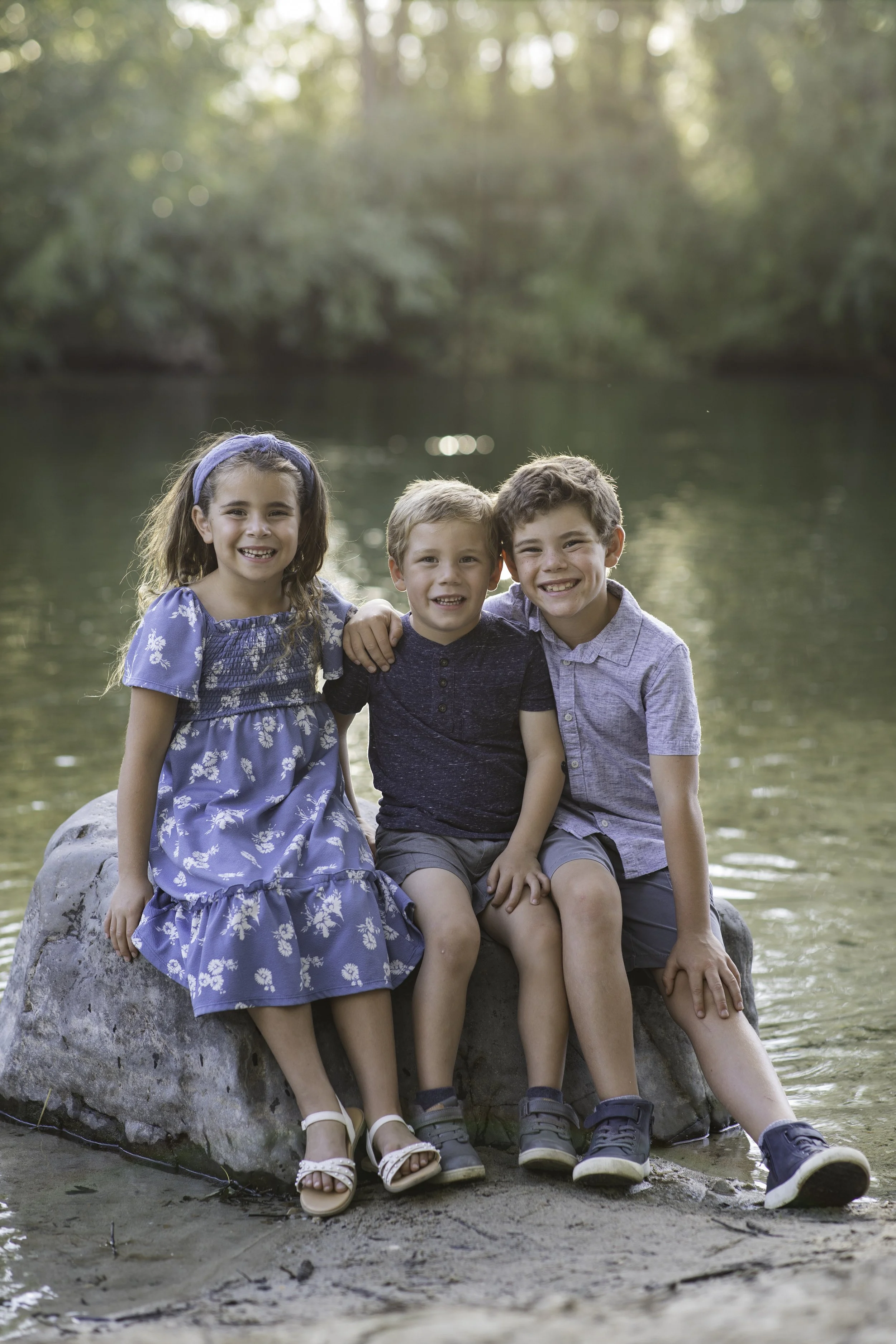 Three children sitting on a large rock by a river, smiling at the camera with a wooded background