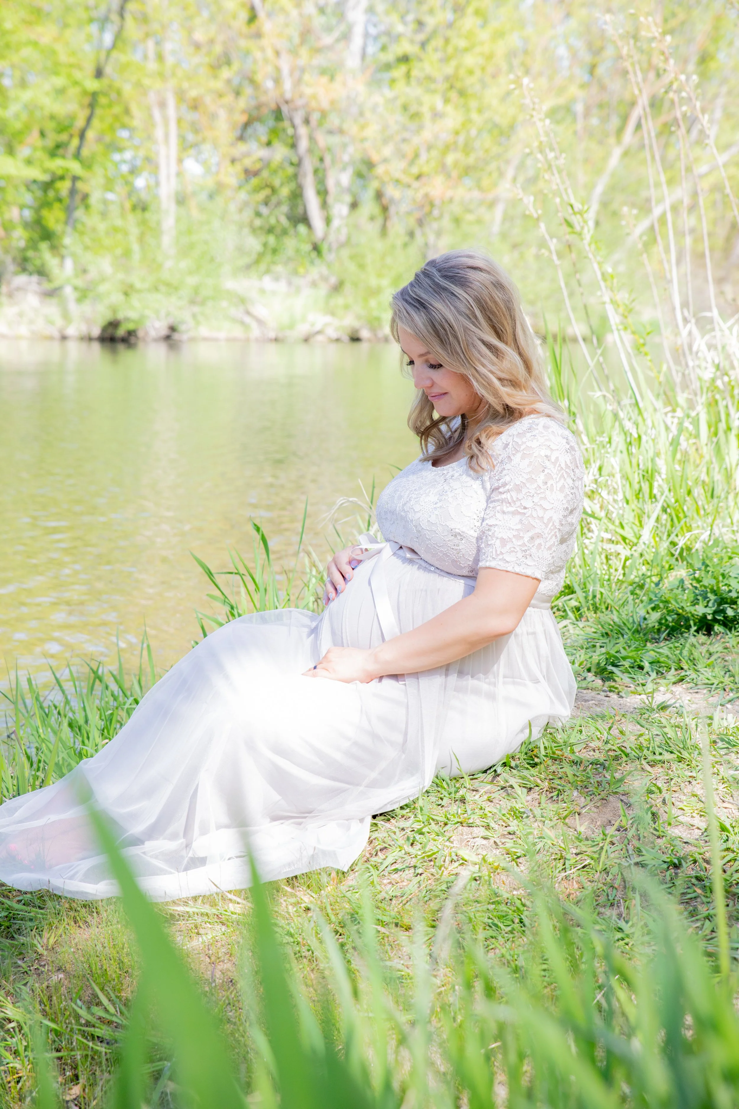 Pregnant woman in a white dress sitting by a riverbank in a lush green outdoor setting.